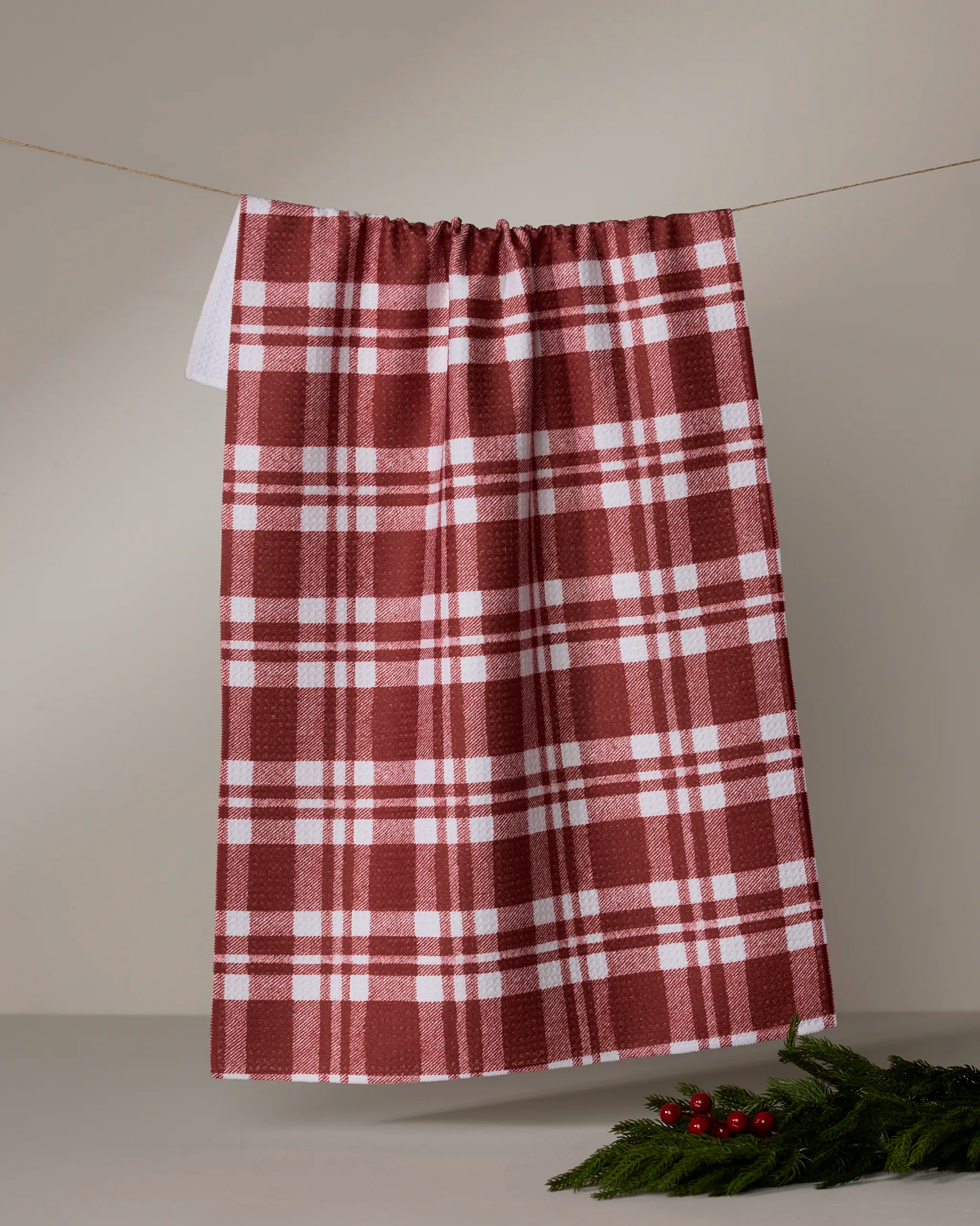 The Geometry Cranleigh dish towel, featuring a red and white plaid design, hangs on a string against a neutral background, accented with a small green pine branch and red berries in the lower right corner.