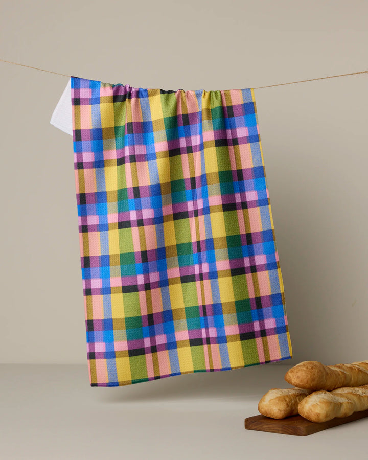The Geometry Crayon tea towel, featuring yellow, blue, green, and pink plaid stripes, hangs above a table with baguettes and a wooden cutting board. Made from quick-drying recycled materials for daily use.