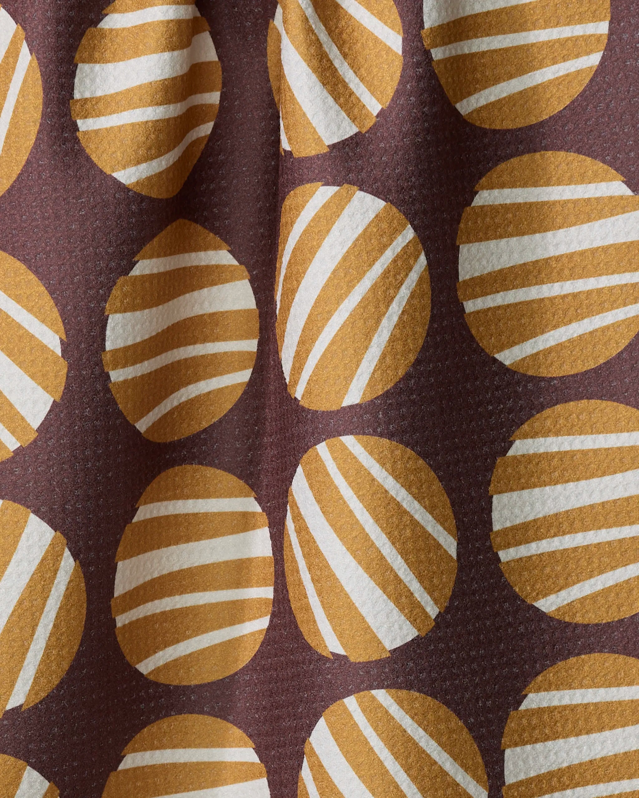 The Crazy Circles tea towel by Lizzie Clark features mustard yellow circles with white and yellow stripes on a dark brown background. Made from slightly textured, quick-drying fabric, it’s perfect for everyday kitchen use.