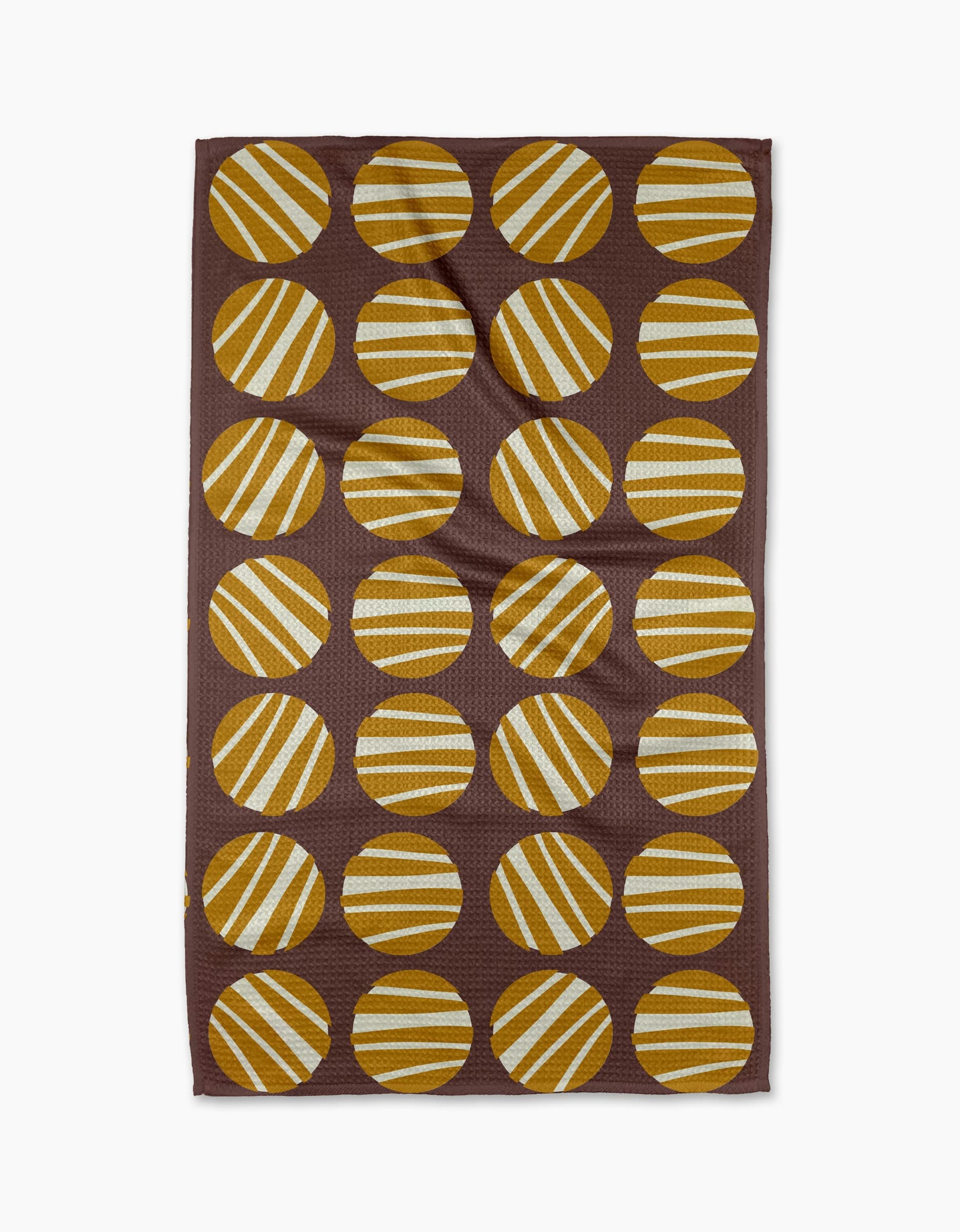 The Crazy Circles tea towel by Lizzie Clark is brown and rectangular, featuring a grid of evenly spaced yellow and white striped circles with diagonal stripes on its ultra-absorbent surface.