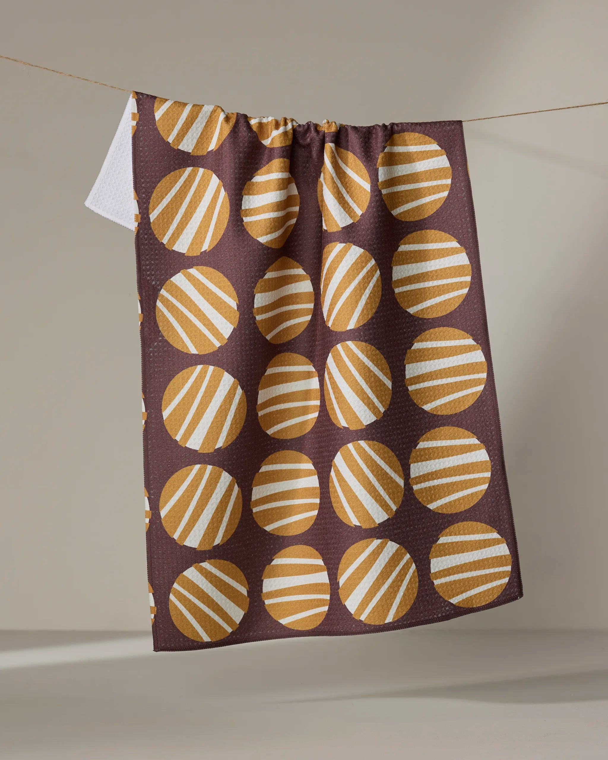 The Crazy Circles tea towel by Lizzie Clark features bold yellow-orange circles with diagonal white stripes on a plain brown, neutral background. Made from recycled materials, it hangs stylishly on a line.