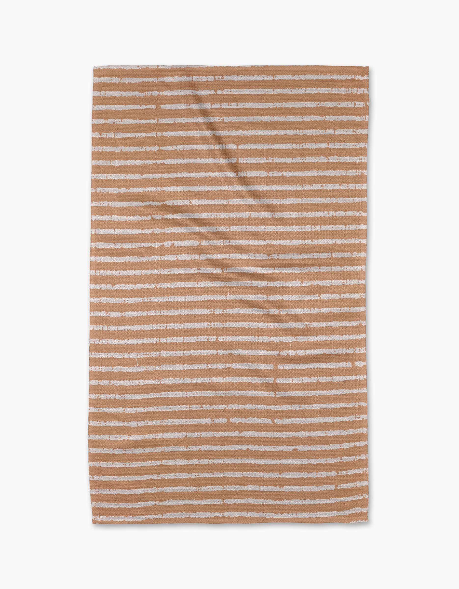 The Geometry Creamsicle Tea Towel, rectangular and made from recycled materials, features horizontal uneven white stripes on a warm tan background and is shown laid flat against a white surface.