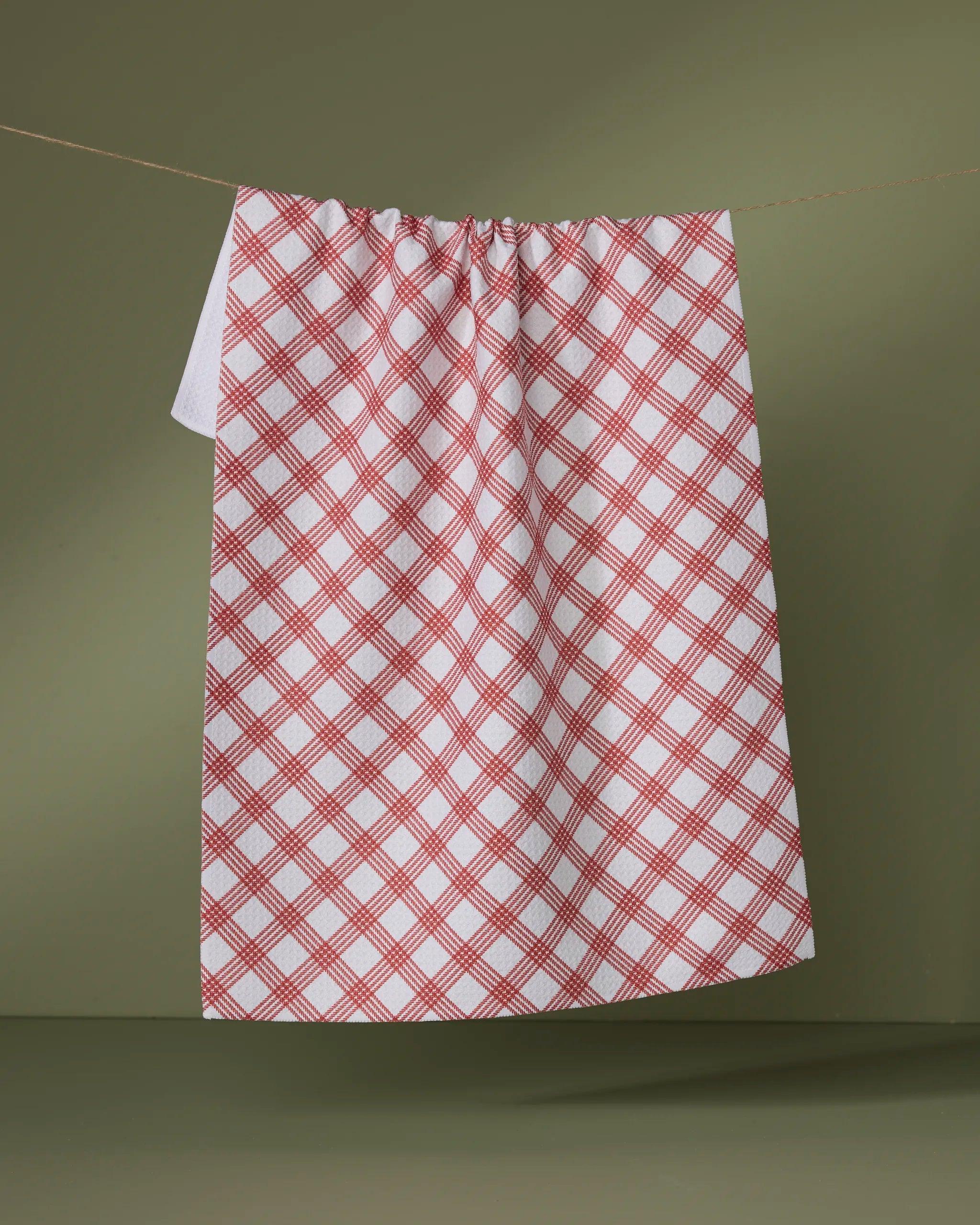 The Crimson Wrap by Geometry, a white tea towel with a red plaid pattern made from recycled materials, hangs on a clothesline against an olive green background.