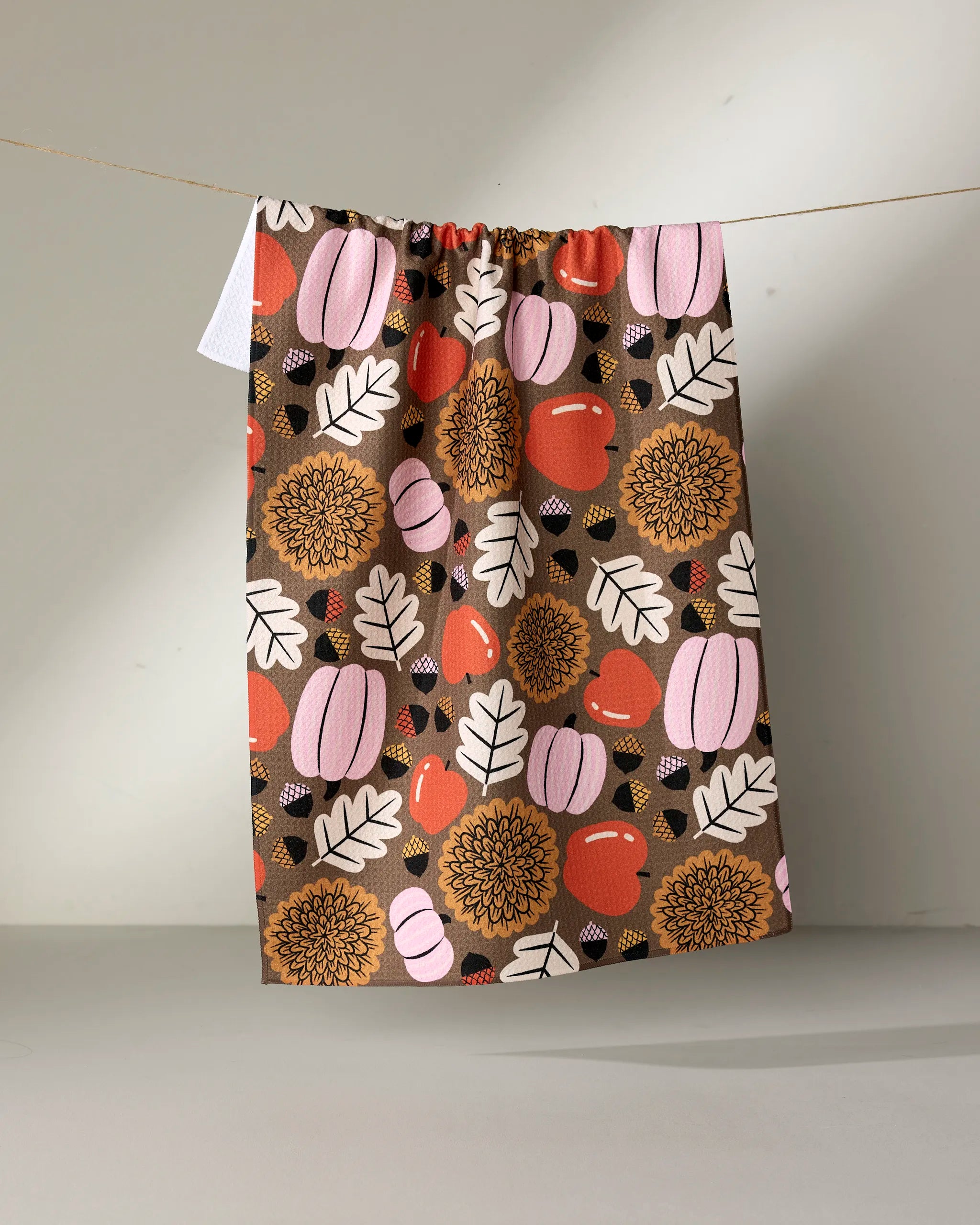The "Crisp and Cozy" tea towel by Megan Roy is made from recycled materials and features a vibrant fall design with pink pumpkins, red apples, yellow flowers, and white leaves.