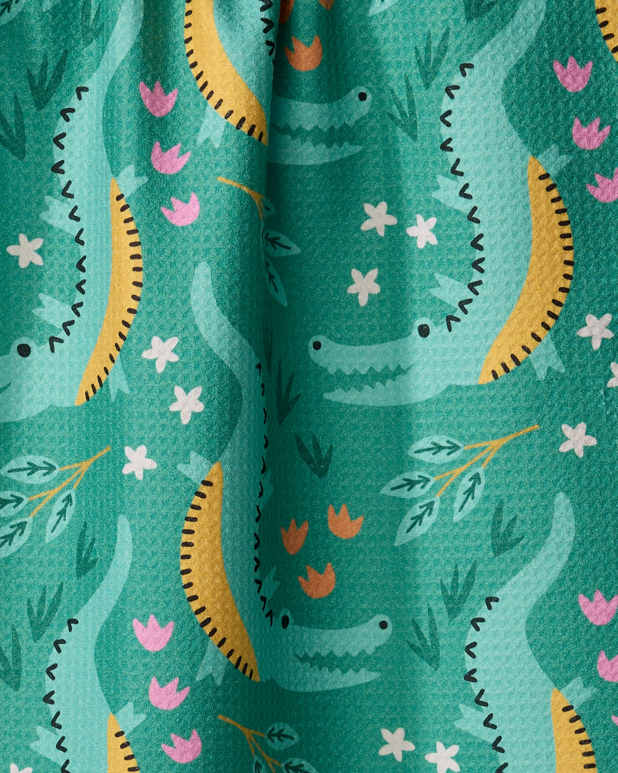 A close-up of the "Crocodiles" recycled kitchen towel by Genna Blackburn, showcasing a whimsical teal design with green crocodiles, yellow bananas, pink and orange flowers, and green leaves.