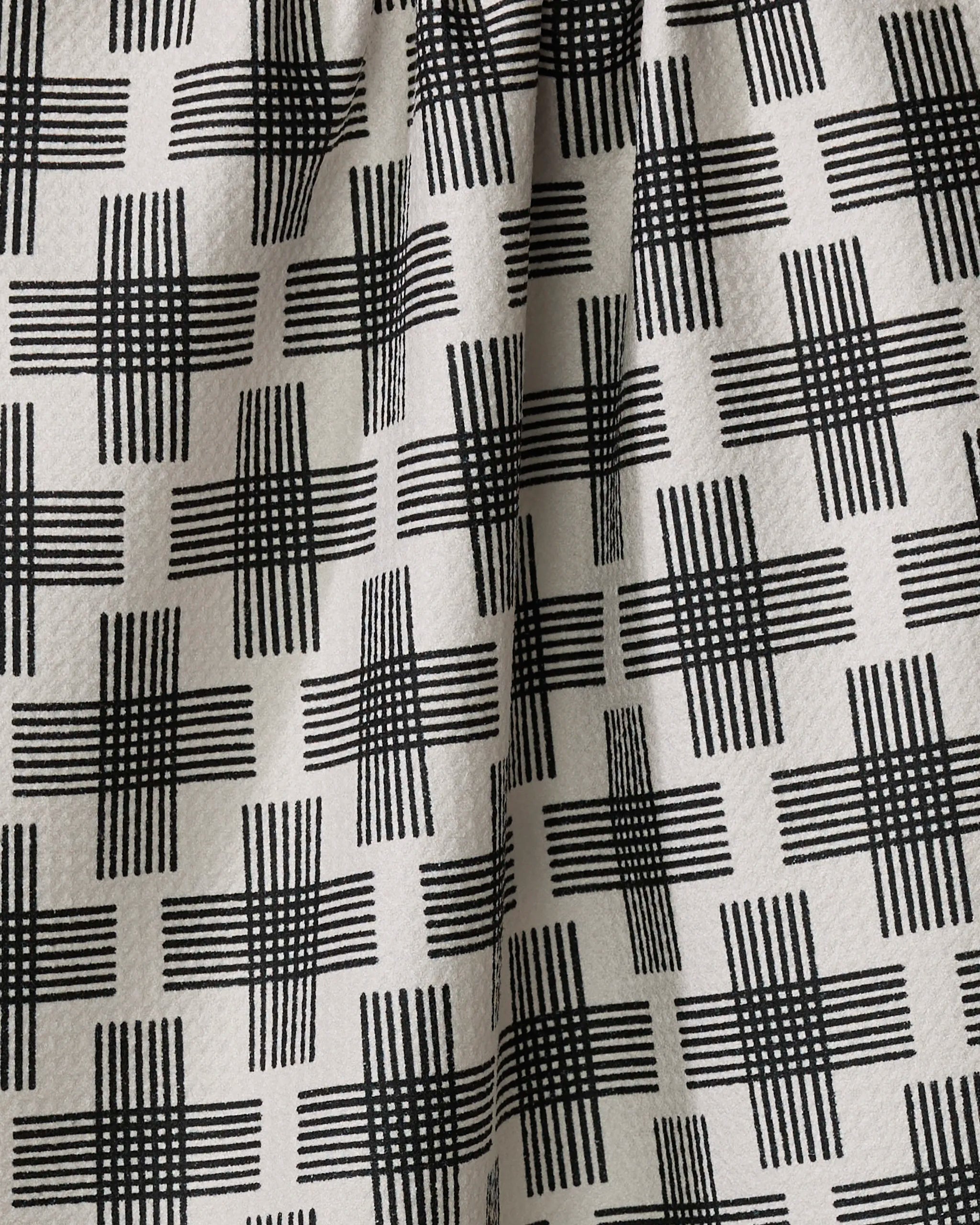 A close-up of the Cross Hatch tea towel by Rosa Kichline displays black geometric crisscross lines on a light background. The ultra-absorbent fabric features gentle folds and a subtly textured surface.