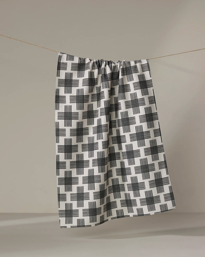 The Cross Hatch tea towel by Rosa Kichline, made from recycled materials and featuring a black-and-white grid pattern, hangs from a string against a neutral background.