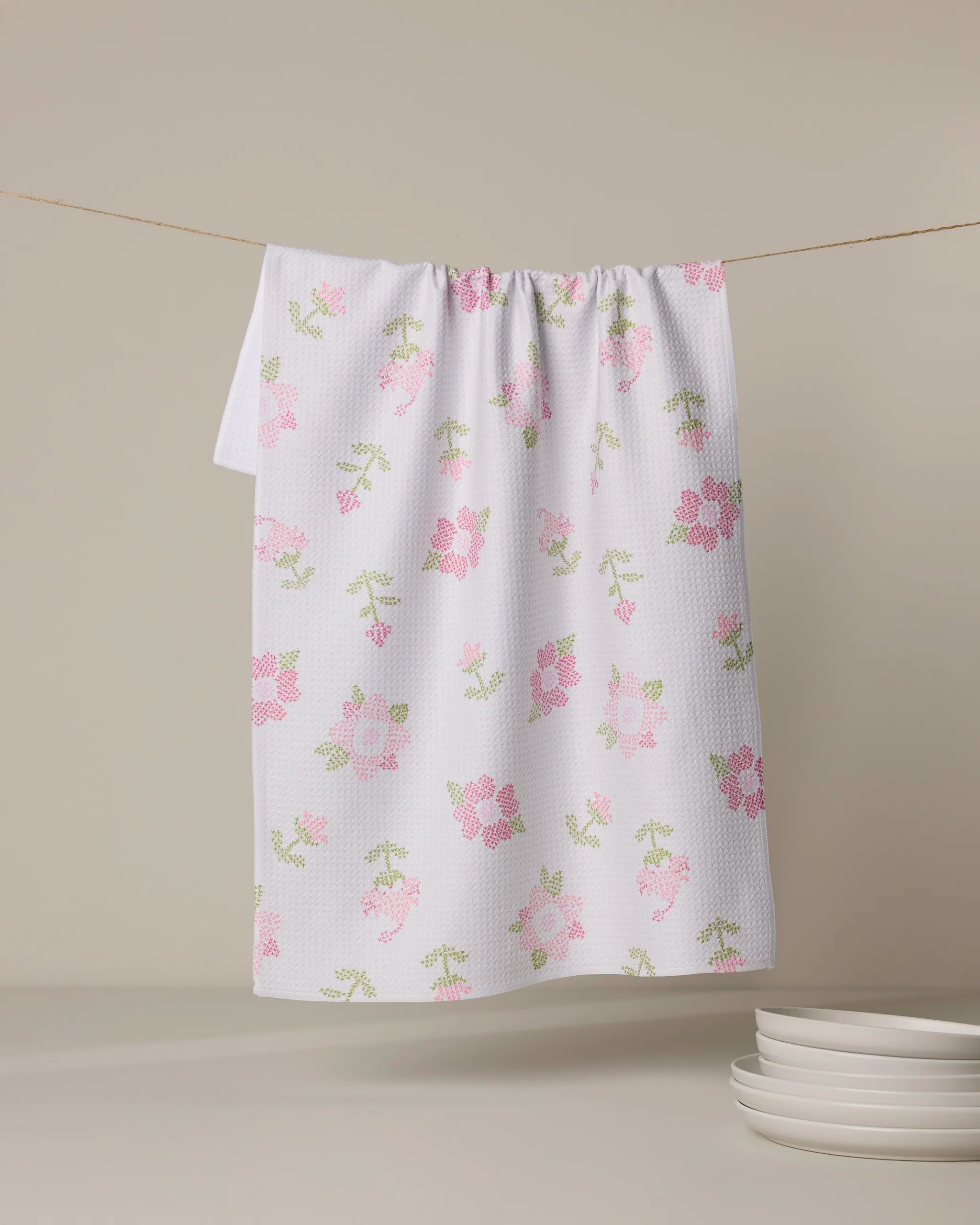 The Geometry Cross Stitch Floral tea towel, featuring a pink and green floral print, hangs on a clothesline above four stacked white plates on a neutral surface. A plain light background highlights the charming kitchen accessory.
