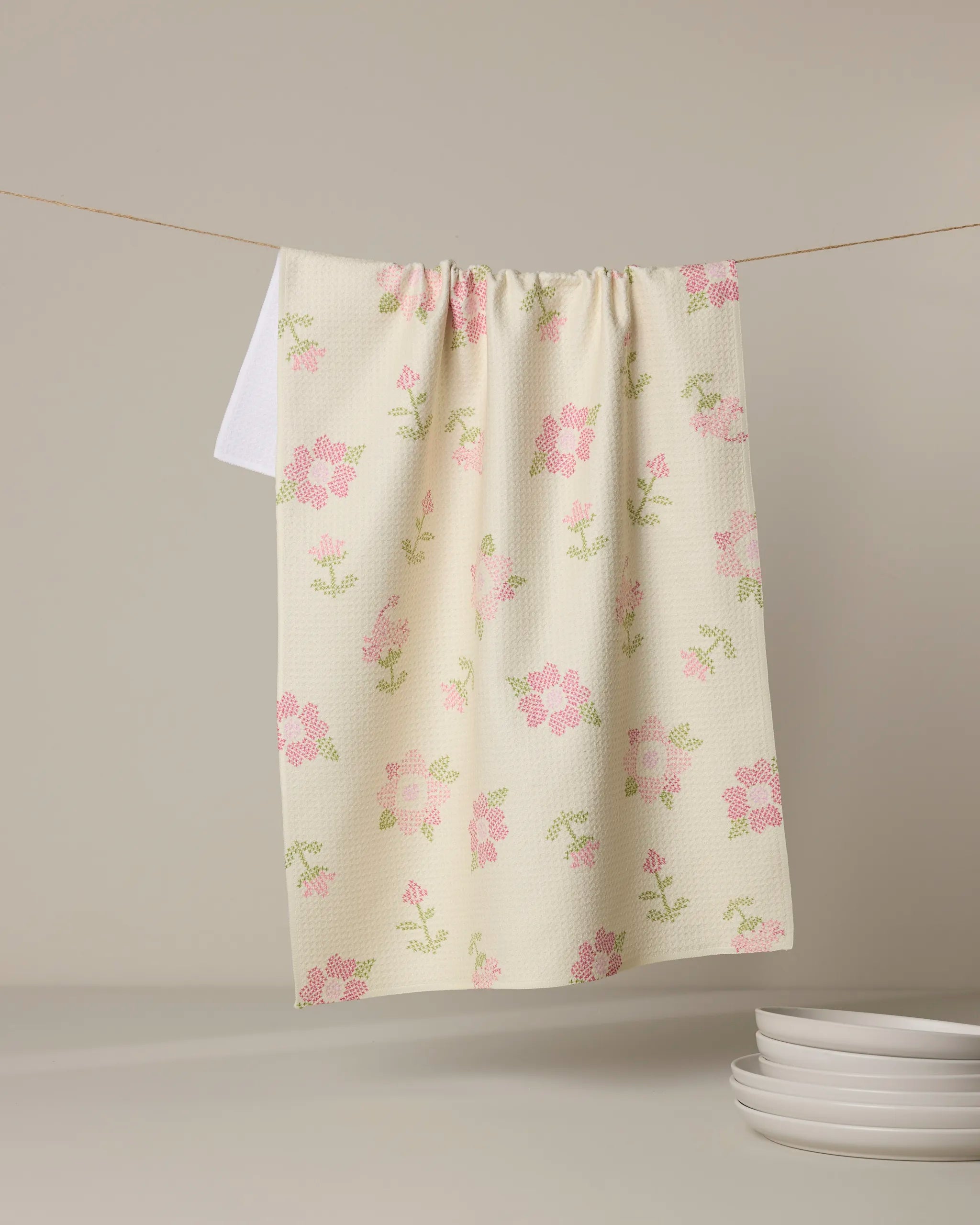 The Geometry Cross Stitch Floral Yellow eco-friendly kitchen towel, featuring a yellow and floral pattern, hangs on a clothesline above white plates against a plain, light background.