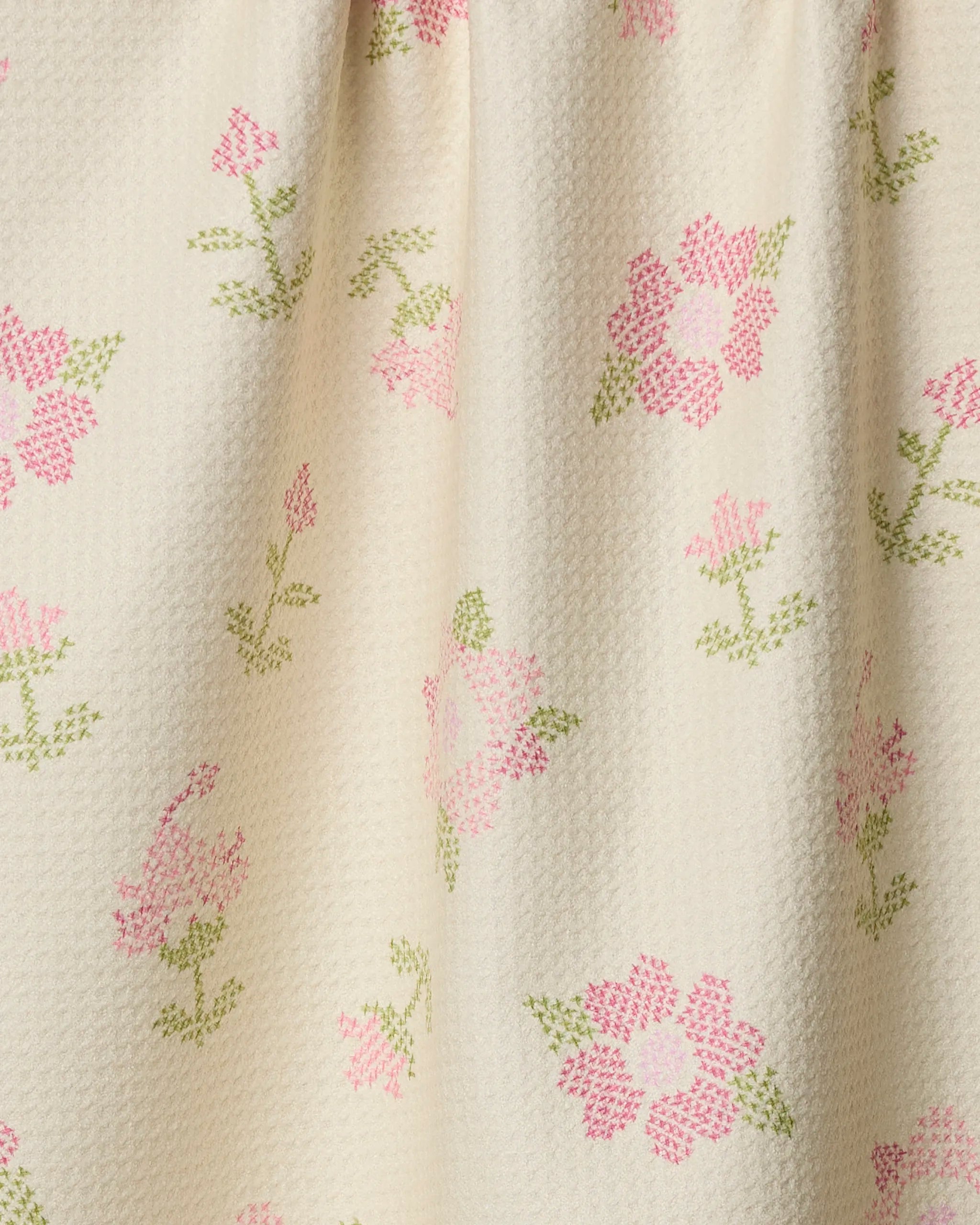 A close-up of cream-colored textured fabric, Geometry's Cross Stitch Floral Yellow kitchen towel showcases pink and green cross-stitched flower patterns for an eco-friendly touch.