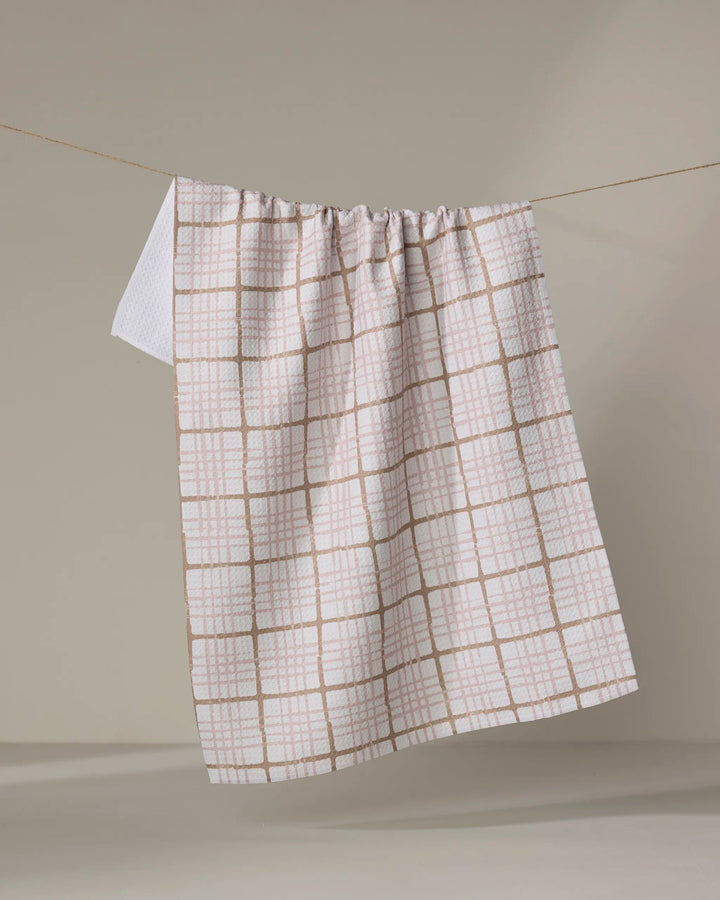 The Studio Jeannot Crossover is a white tea towel with a light brown grid, hanging over a clothesline against a beige background. Made from ultra-absorbent, recycled materials, it’s stylish and eco-friendly.