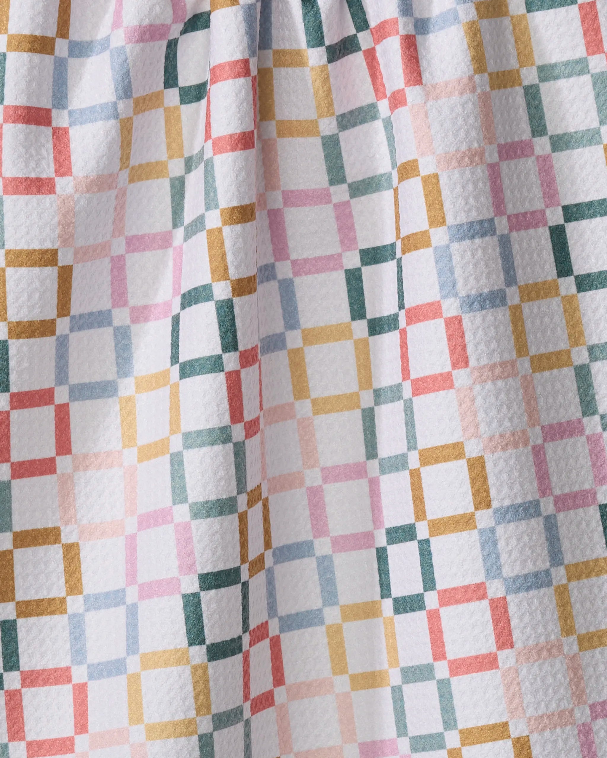 A close-up of the Crossroads kitchen towel by Charlie Rowan Designs shows its softly textured, gathered fabric made from recycled materials. The white background features a colorful grid pattern with green, pink, yellow, and red lines.