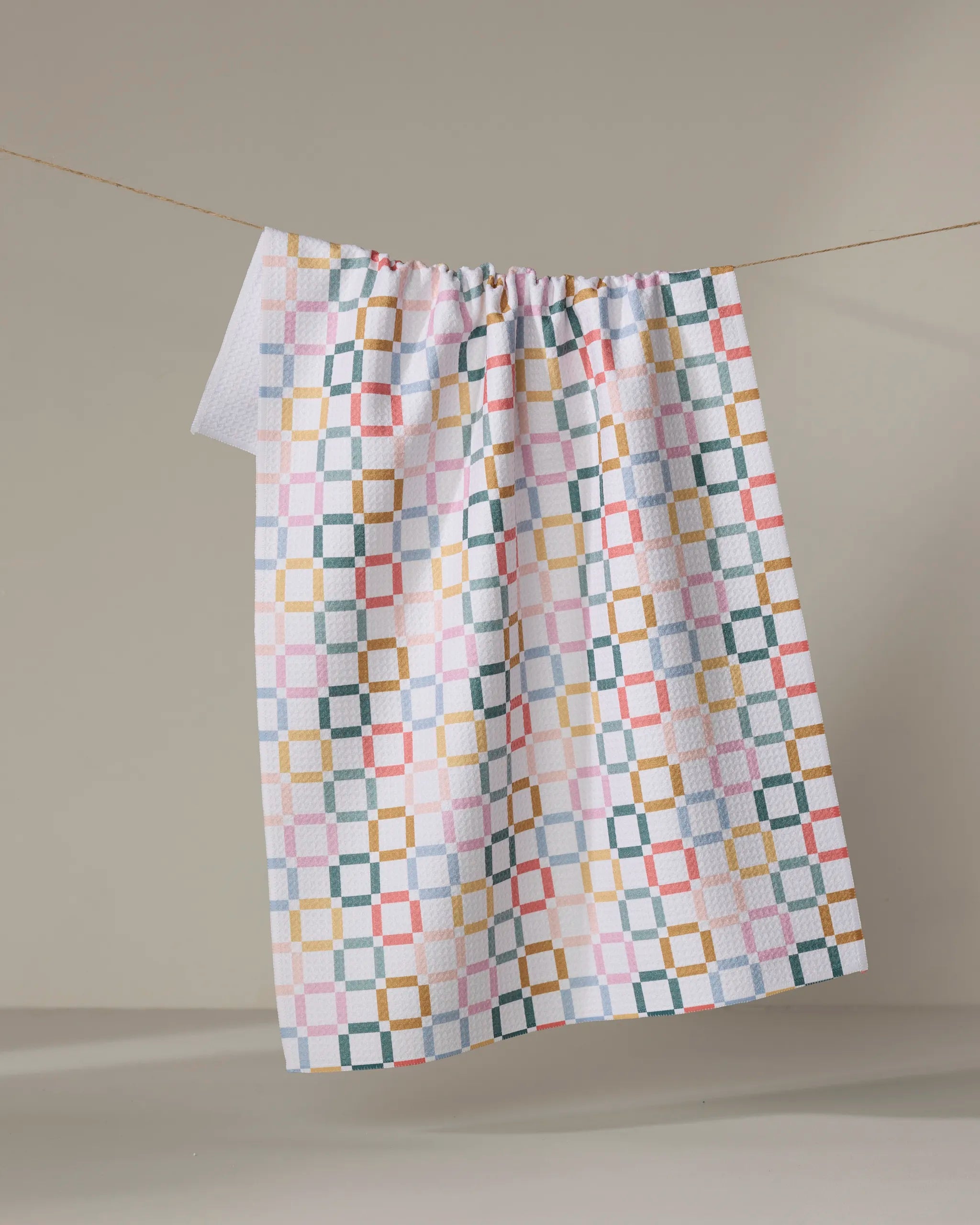 The "Crossroads" kitchen towel by Charlie Rowan Designs, featuring a colorful geometric square and grid pattern, hangs from a line against a plain beige background.