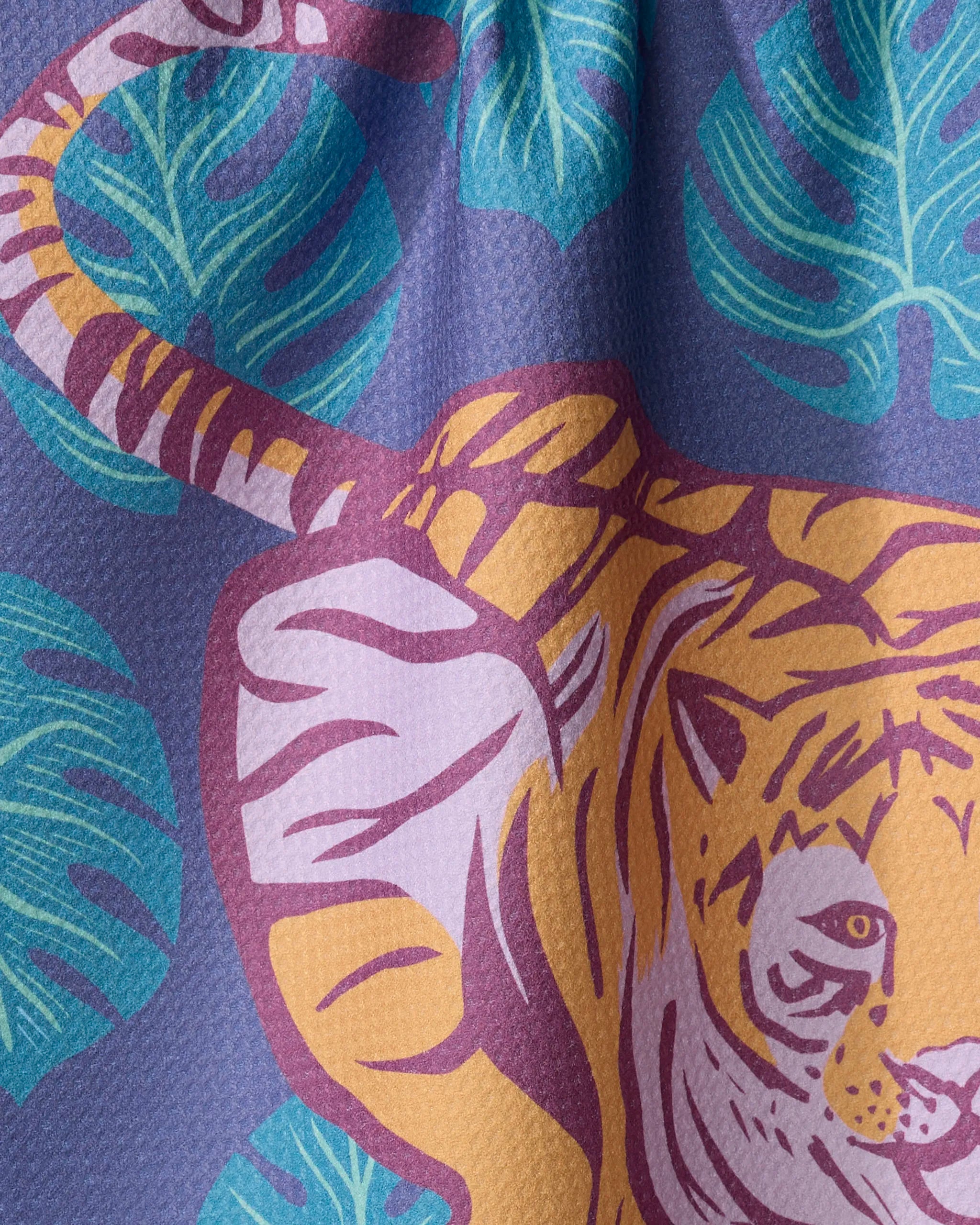 The Crouching Cat tea towel by Elisabeth Wing features a vibrant cat peeking from large teal leaves on a textured purple background. Made from recycled materials, its orange and white fur pops with pink details and a striped tail.