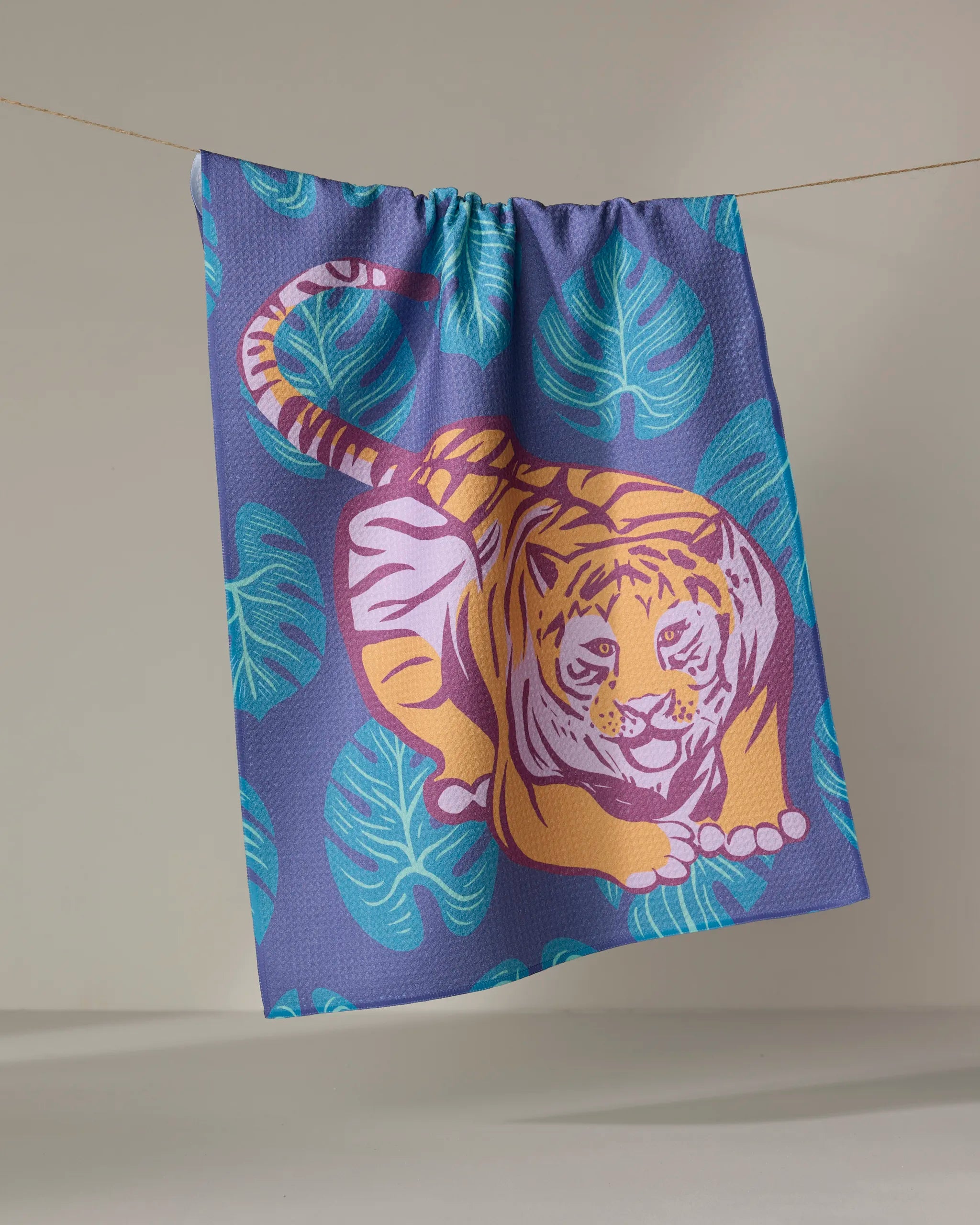 The "Crouching Cat" tea towel by Elisabeth Wing, made from recycled materials, displays a vibrant cat with blue tropical leaves on purple, hanging against a neutral backdrop.