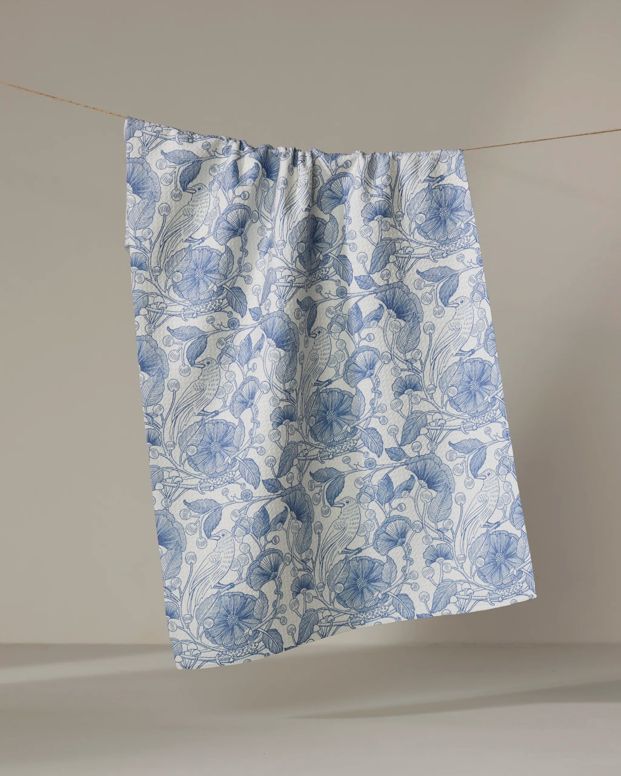 The Crowberries tea towel by Katie O'Shea, featuring a light blue floral pattern, hangs from a thin string against a plain beige background.
