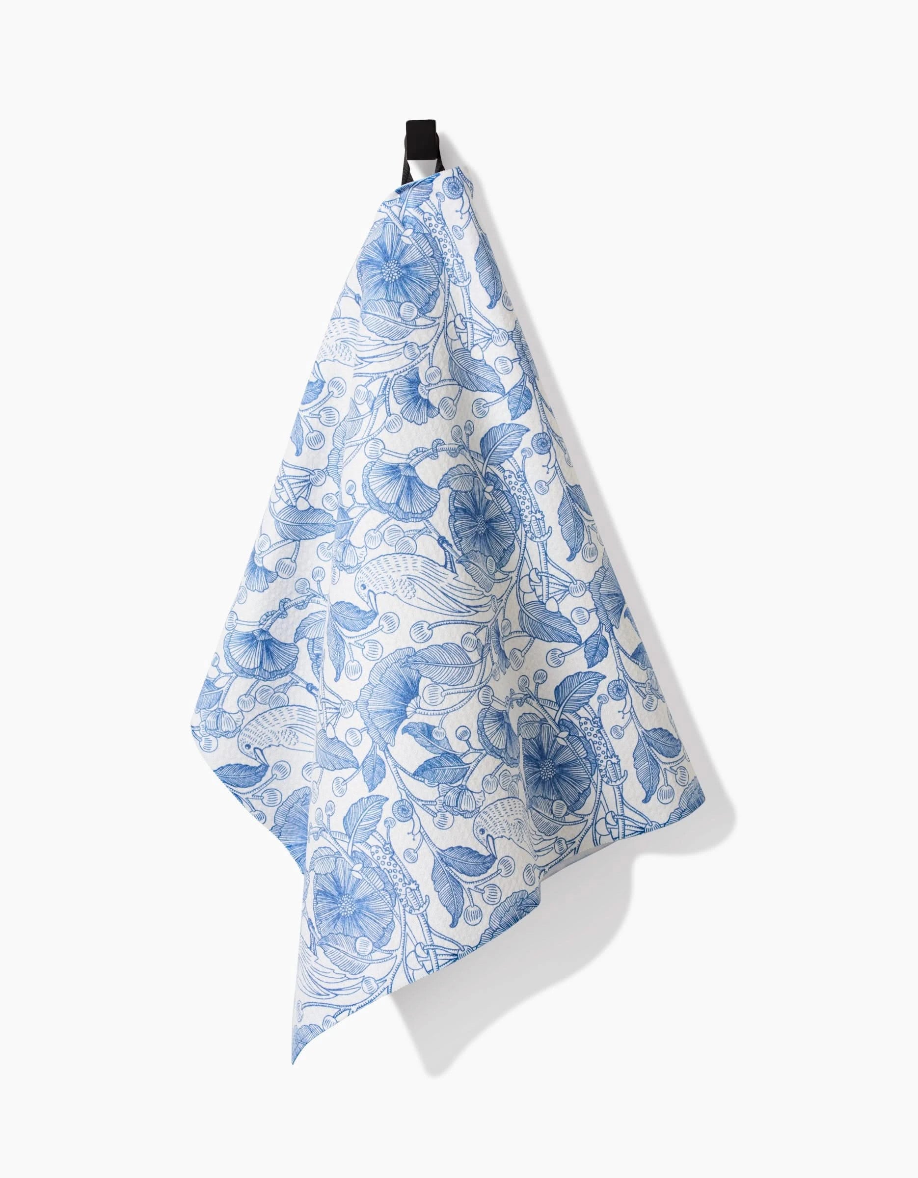 A Katie OShea Crowberries tea towel, ultra-absorbent and decorated with a blue floral design, hangs from a black hook against a plain white background, casting a soft shadow on the wall.