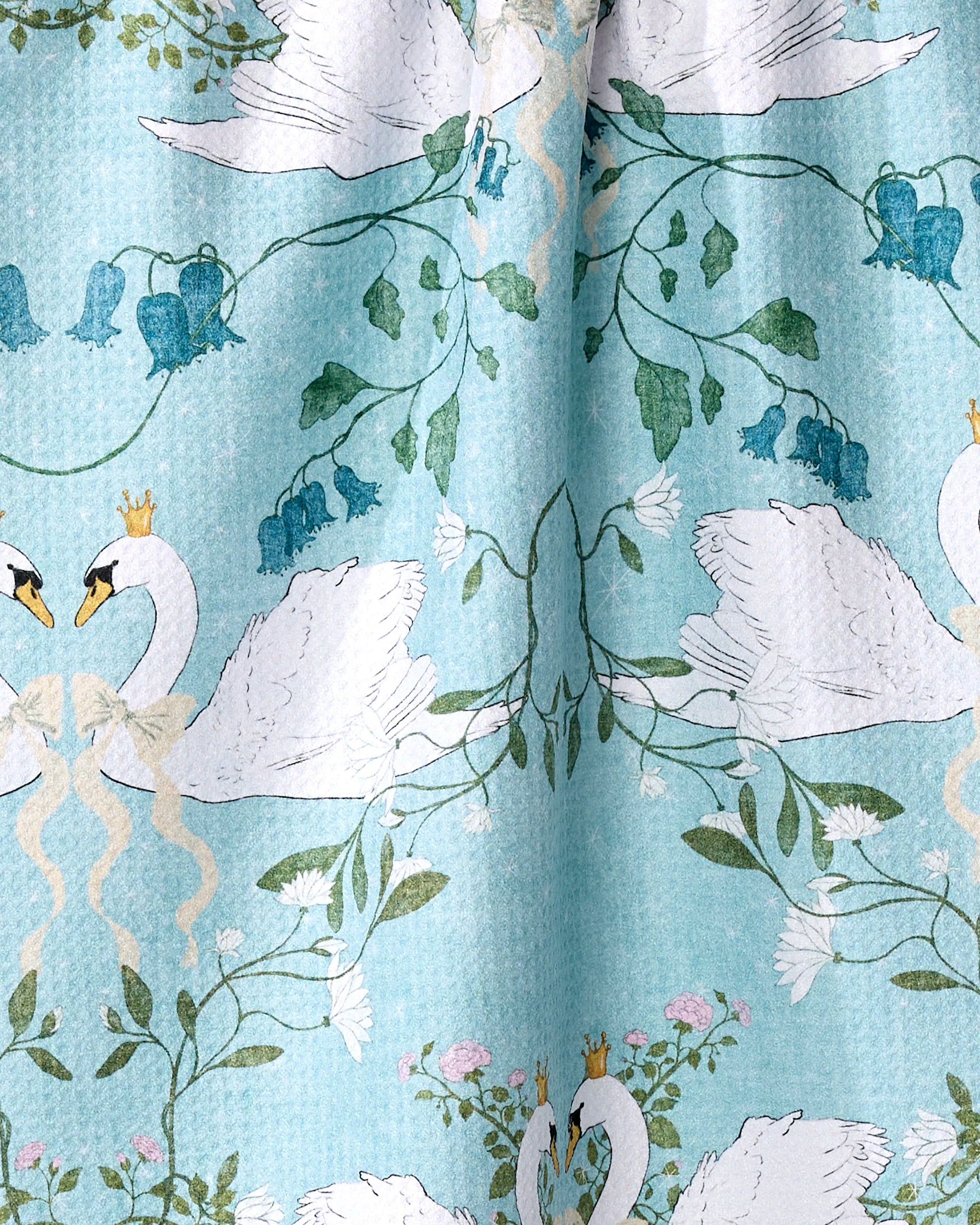 The Pascale Dilger "Crowned Grace" tea towel is ultra-absorbent, made from recycled materials, and features white swans with gold crowns, green vines, and blue flowers on a light blue background. Some swans form heart shapes with their necks.