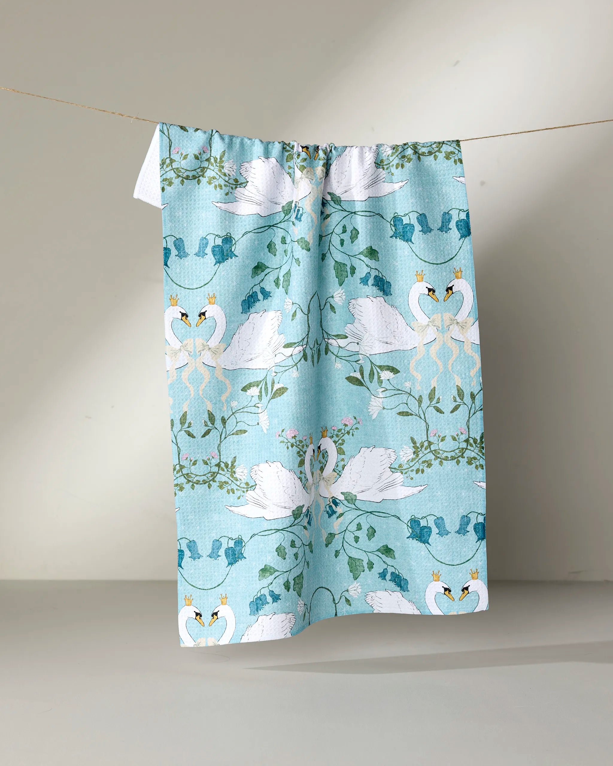 The Crowned Grace tea towel by Pascale Dilger is blue, made from recycled materials, and features white swans forming hearts amid green leaves, pink flowers, and blue crowns, displayed on a clothesline against a neutral background.