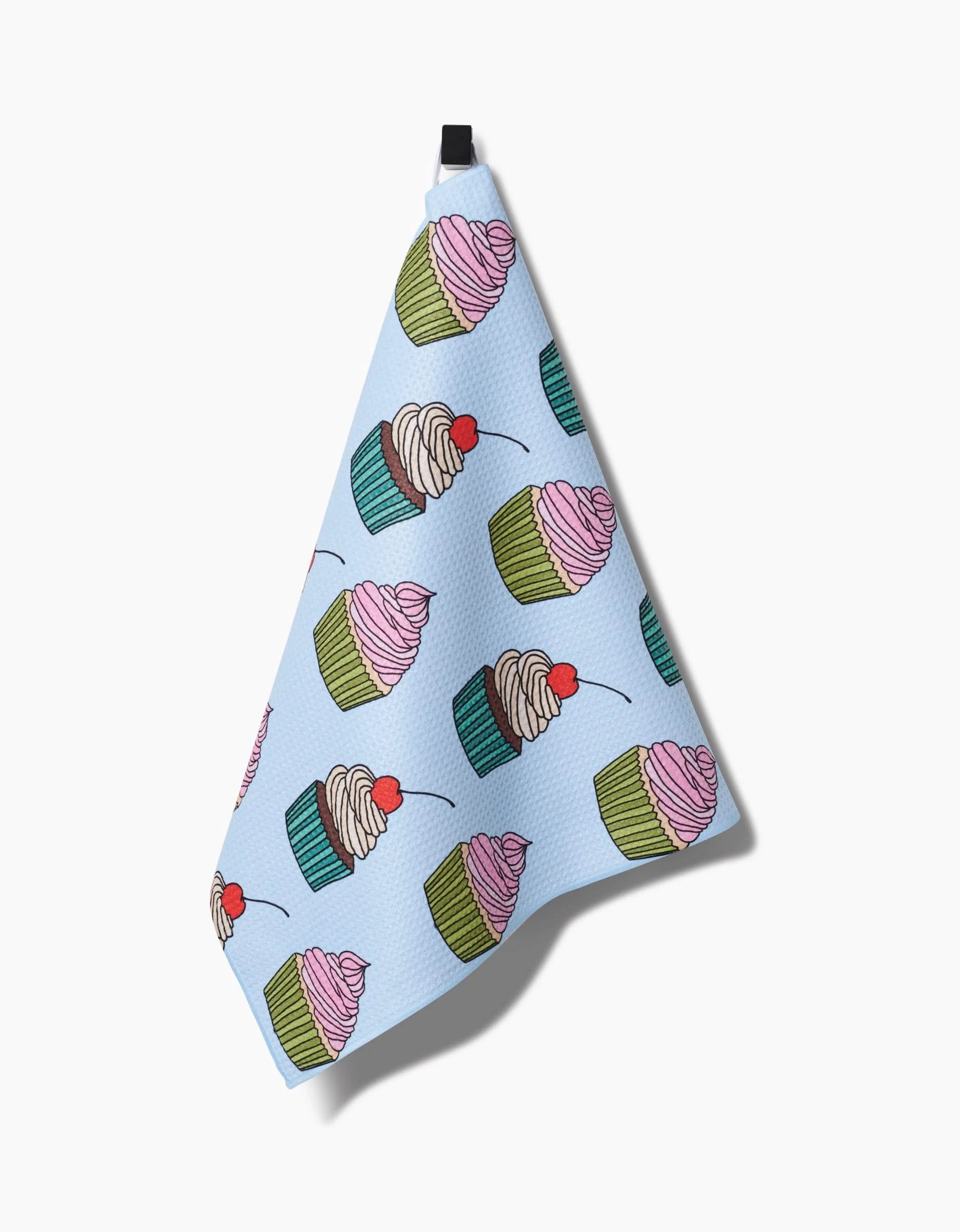 The Cupcake Love tea towel by Amy Droubay is blue, made from recycled materials, and features a colorful cupcake pattern with pink and white frosting plus a cherry. It hangs from a black loop against a white background.