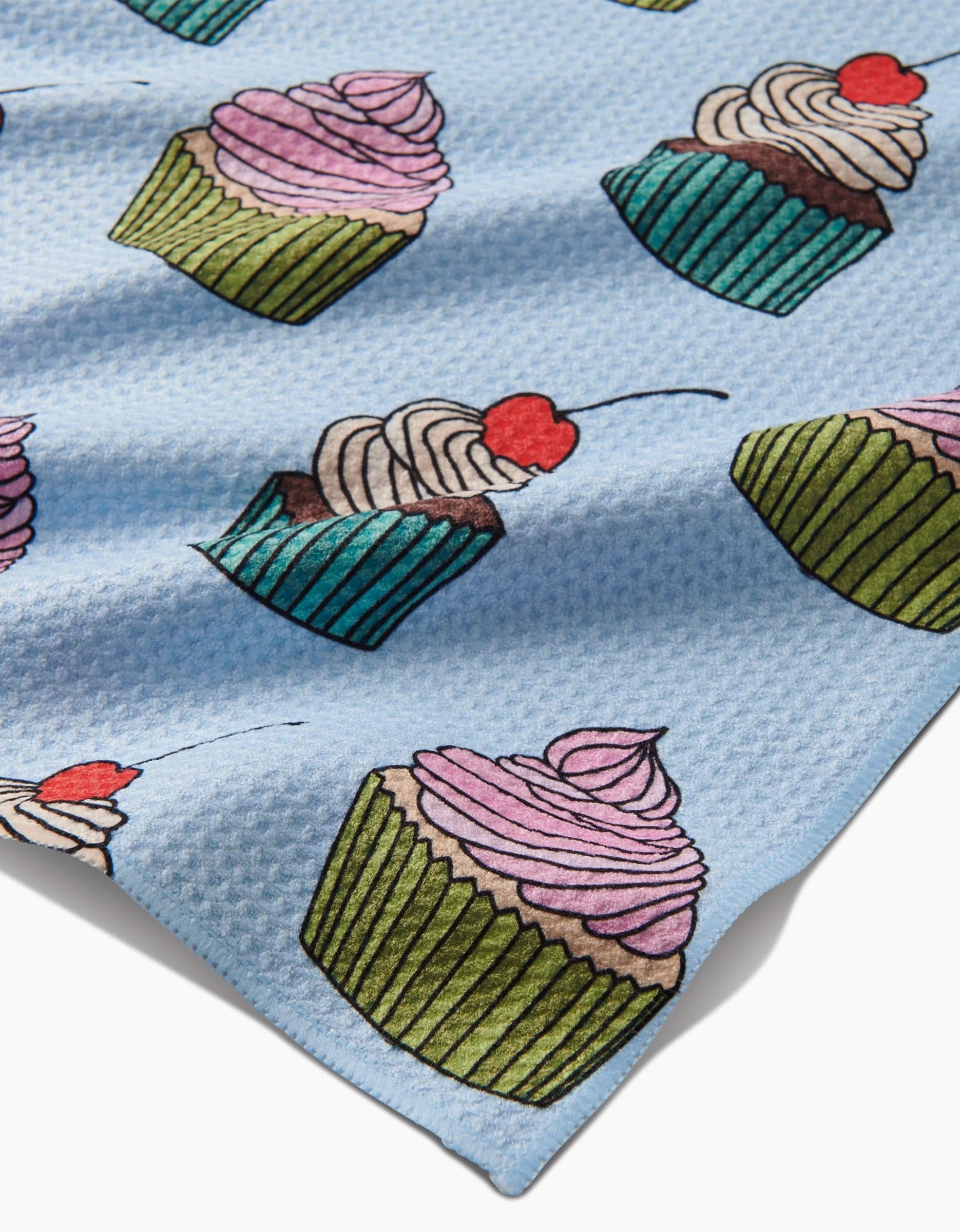 Cupcake Love by Amy Droubay: a light blue textured Geoweave tea towel made from recycled materials, featuring illustrated cupcakes with green or blue wrappers, pink or white frosting, and red cherries.