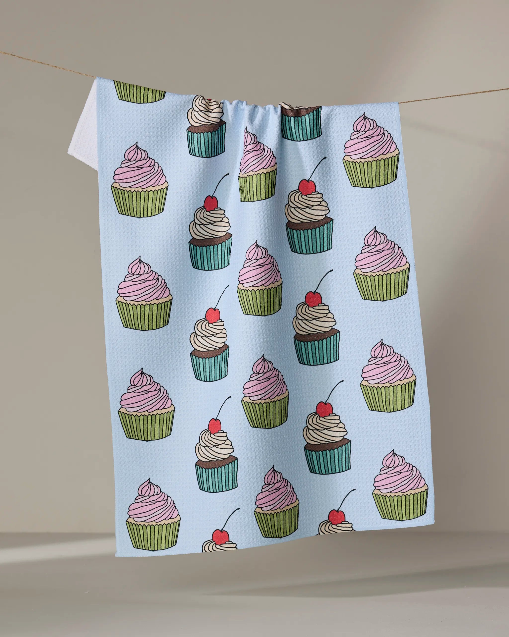 The "Cupcake Love" tea towel by Amy Droubay, crafted from Geoweave fabric, showcases colorful cupcakes with swirled frosting and cherries, hanging on a clothesline against a neutral backdrop.