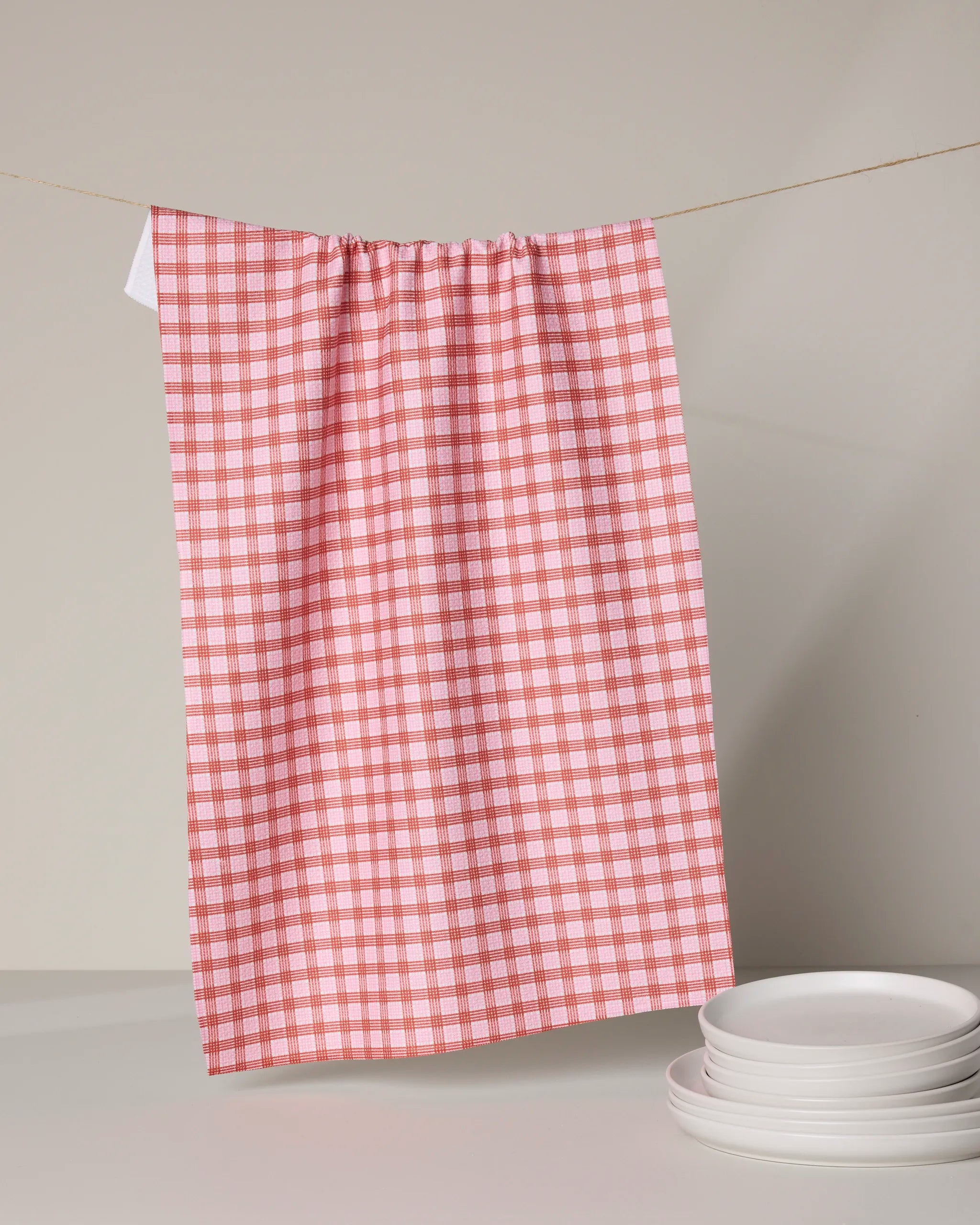 The Geometry Cupids Plaid tea towel, featuring a red and white checkered design, hangs on a string above a white surface with a stack of white plates in the corner. The softly lit background creates a neutral setting.