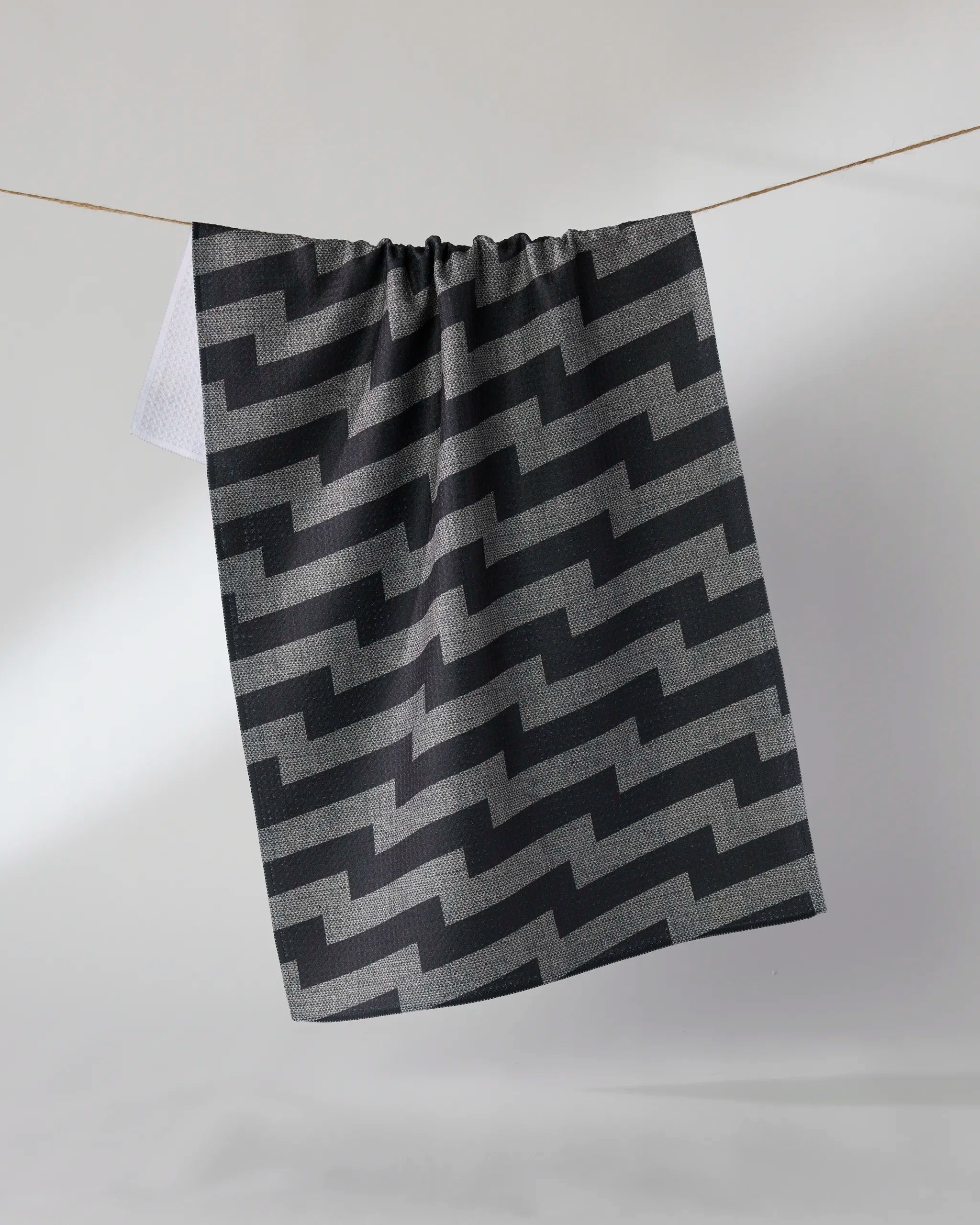 A dark gray, ultra-absorbent Geometry "Current" towel with a bold black zigzag pattern hangs on a clothesline against a plain light background.