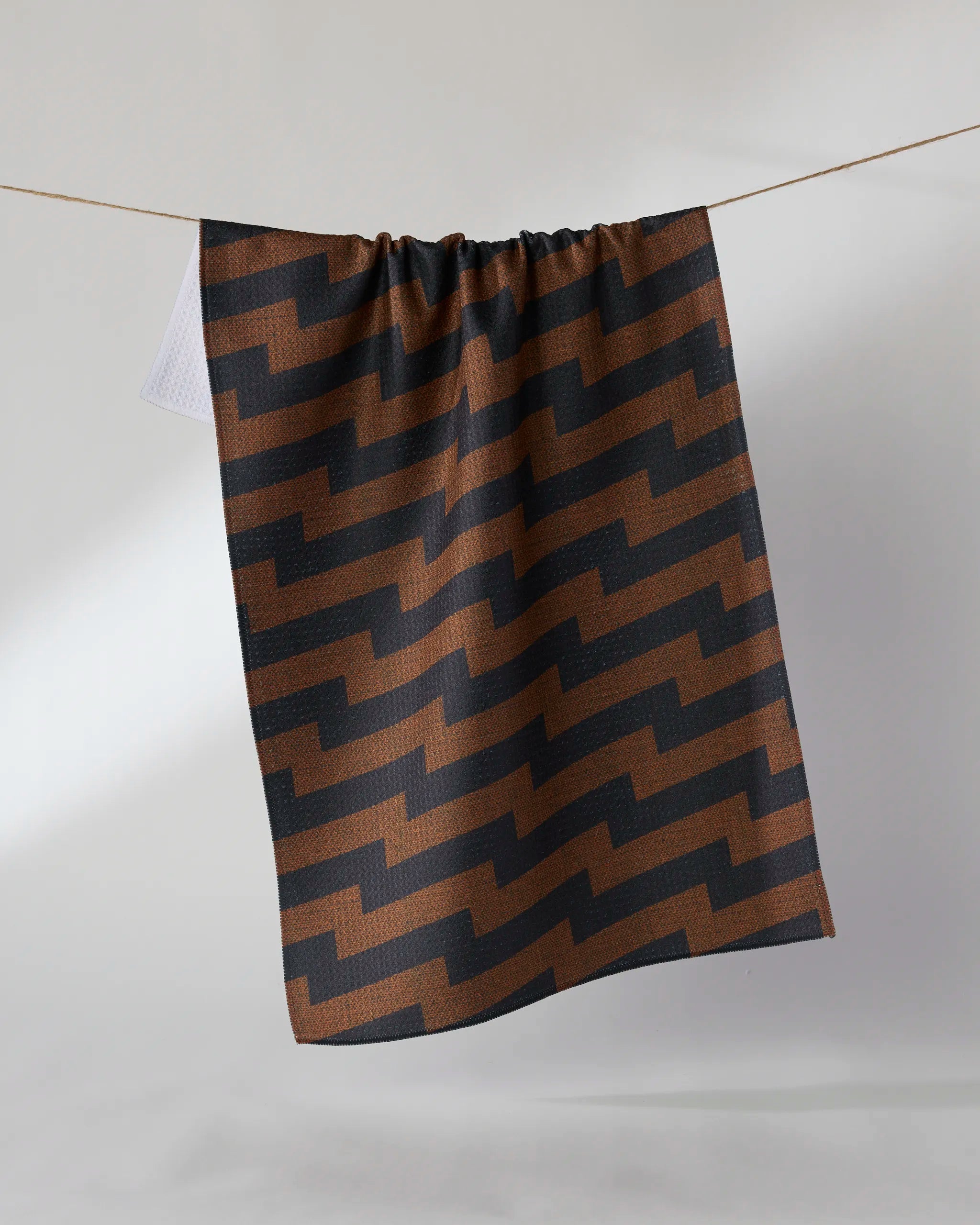 The Geometry "Current" tea towel, featuring a brown and black geometric zigzag pattern, hangs on a line against a simple, light background.