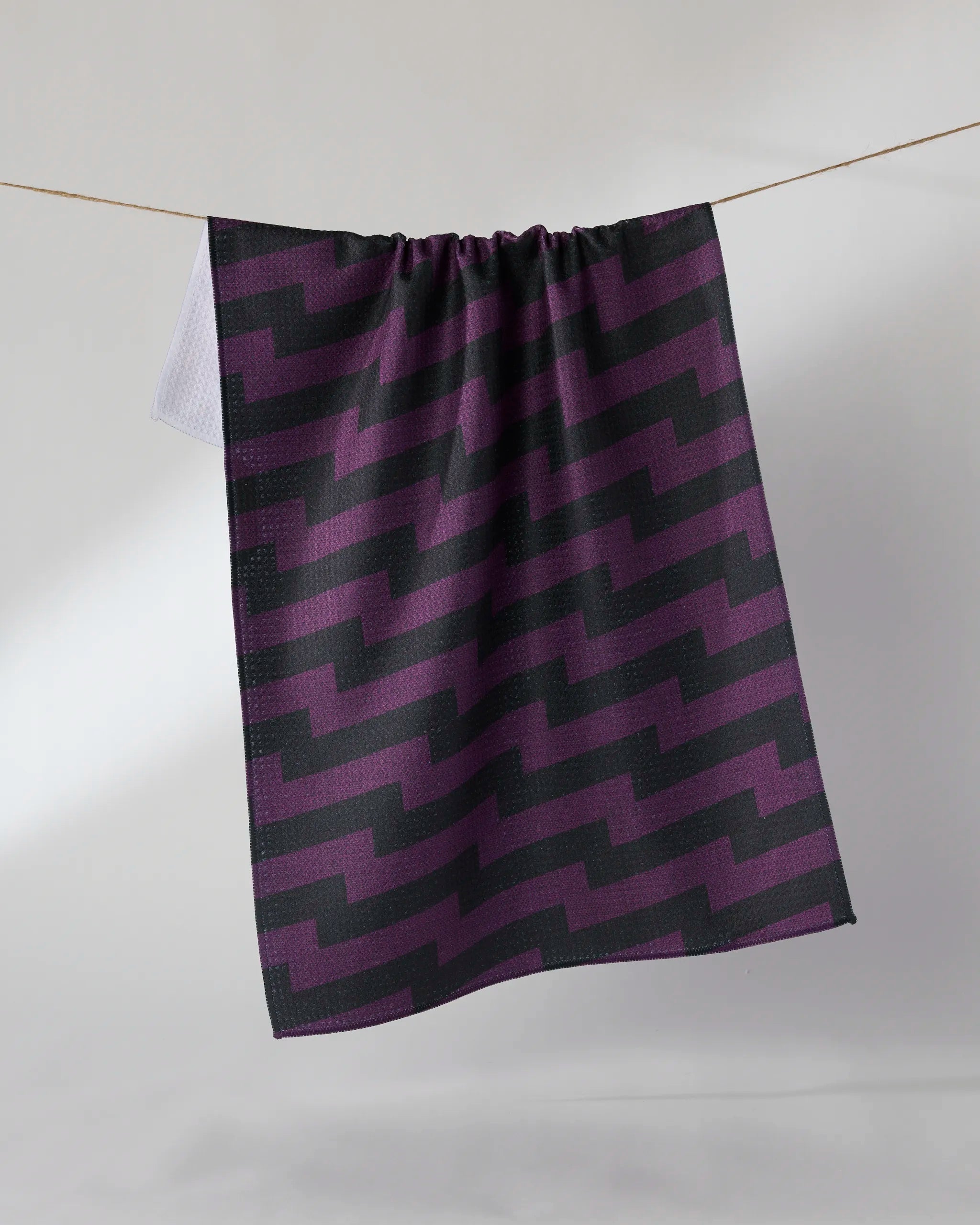The Geometry "Current" tea towel in dark purple and black zigzags hangs on a clothesline against a plain light background.