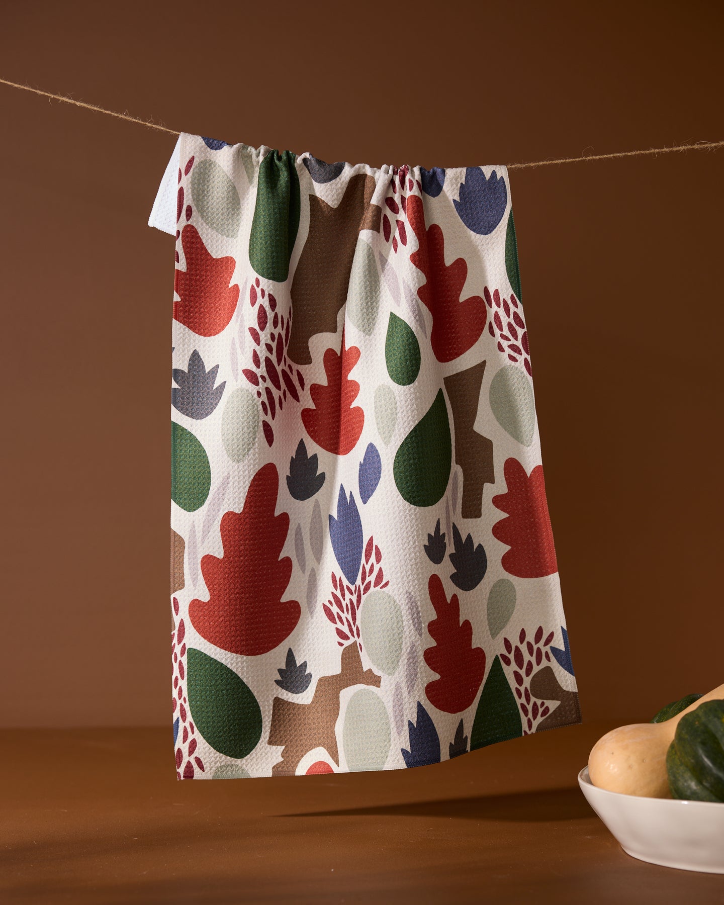 A Geometry Cut Paper Leaves Cream tea towel with a colorful leaf pattern hangs on a string. In the bottom right, part of a plate with potatoes and green squash appears against a brown background.