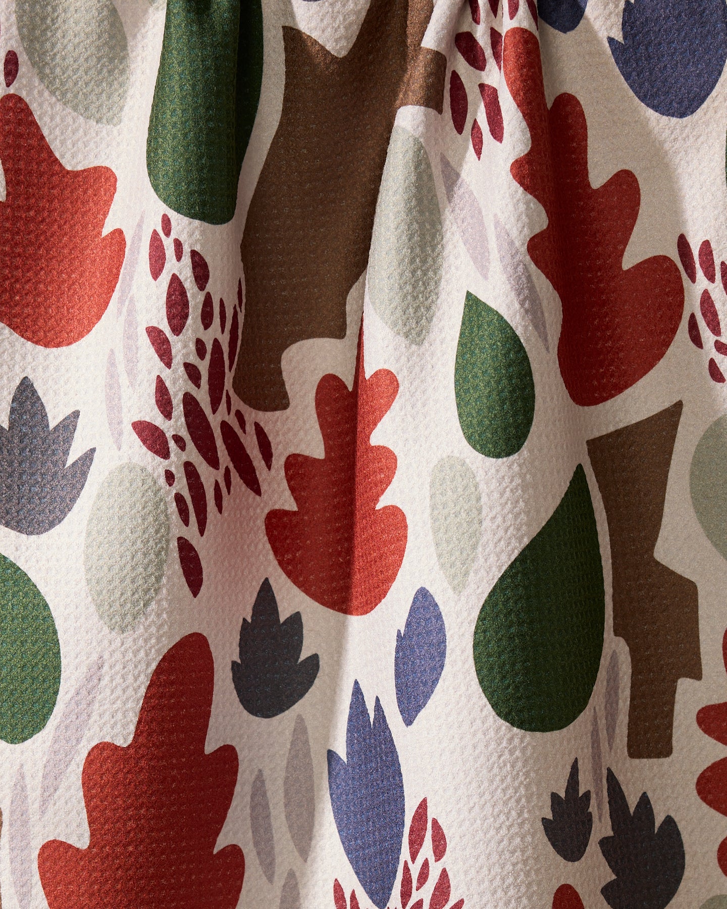 A close-up of the Geometry Cut Paper Leaves Cream tea towel, showcasing a colorful abstract leaf pattern in red, green, brown, blue, and gray on white. The ultra-absorbent towel is shown slightly wrinkled or draped.