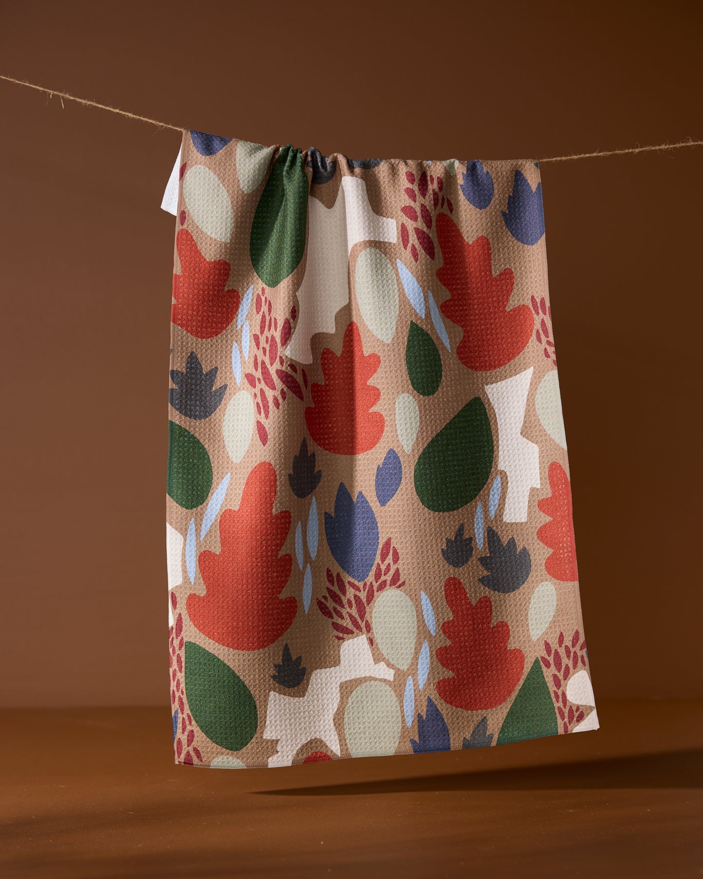 The Geometry Cut Paper Leaves Mocha tea towel, featuring abstract red, green, navy, and beige foliage shapes, hangs on a string against a plain brown background.
