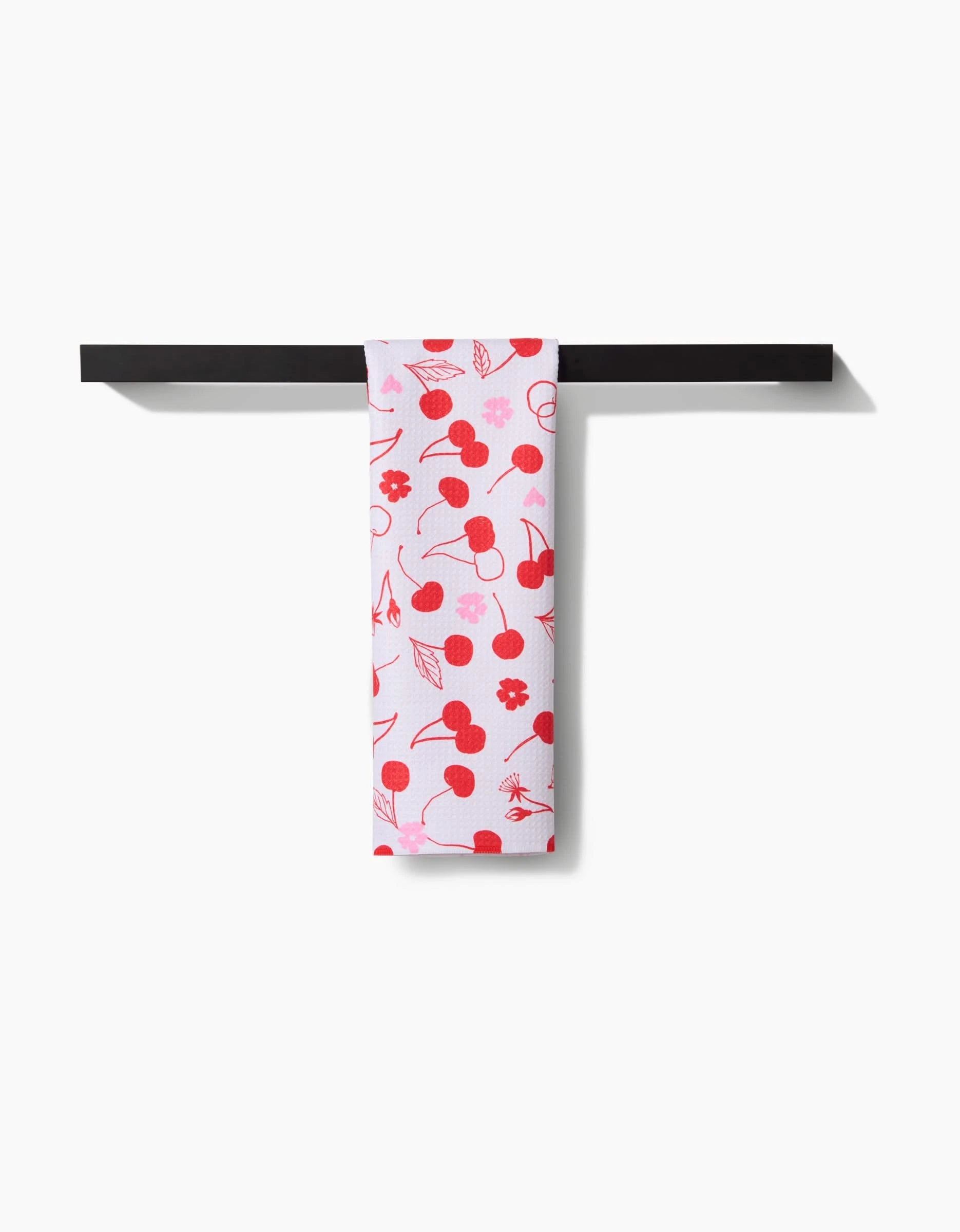 The Elana Gabrielle Cute Cherry white recycled kitchen towel, featuring cherry and flower patterns, hangs neatly over a horizontal black bar against a plain white background.