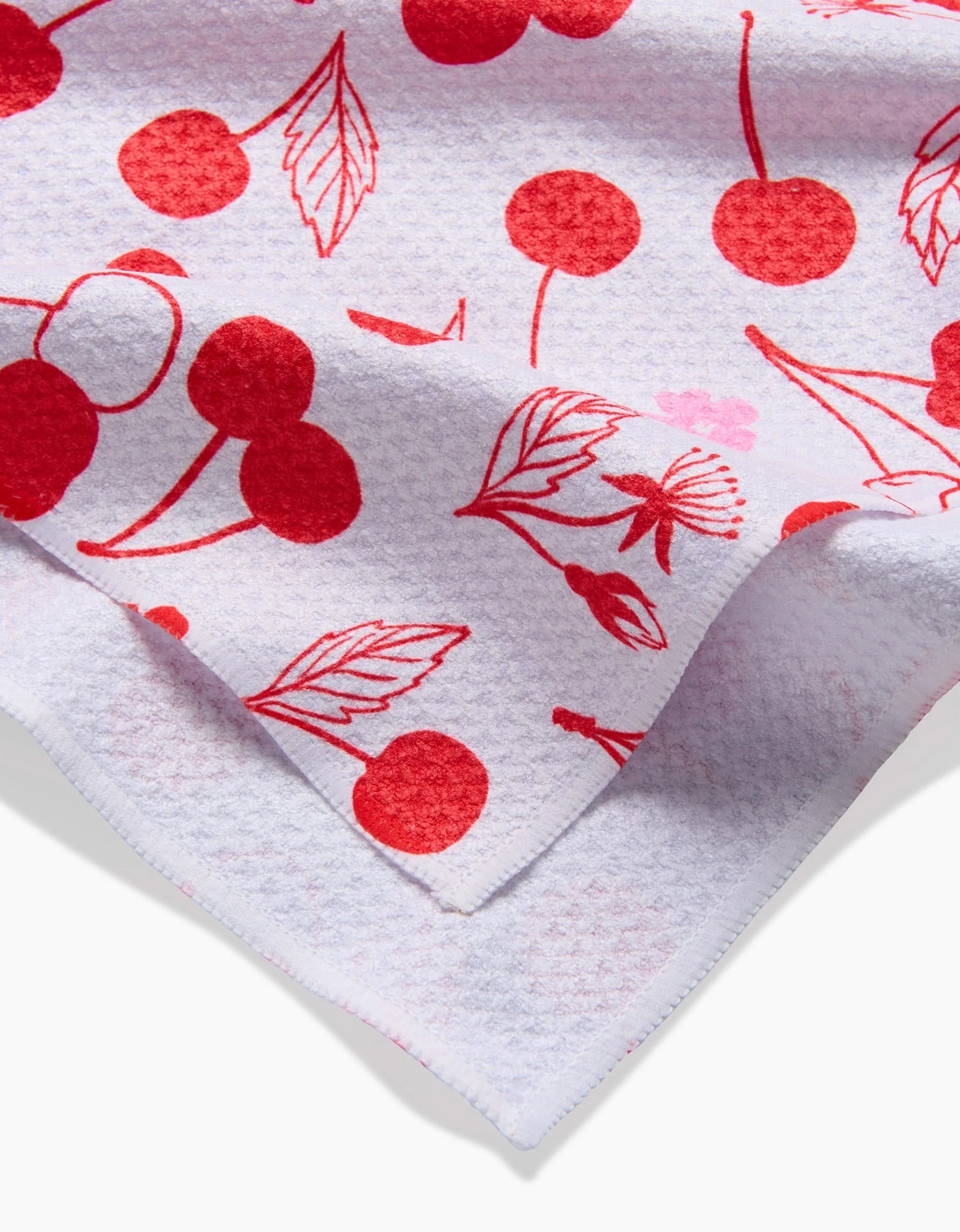 A close-up of Elana Gabrielles Cute Cherry tea towel, showcasing its white textured fabric with a red cherry and leaf design, and a folded corner revealing the underside.