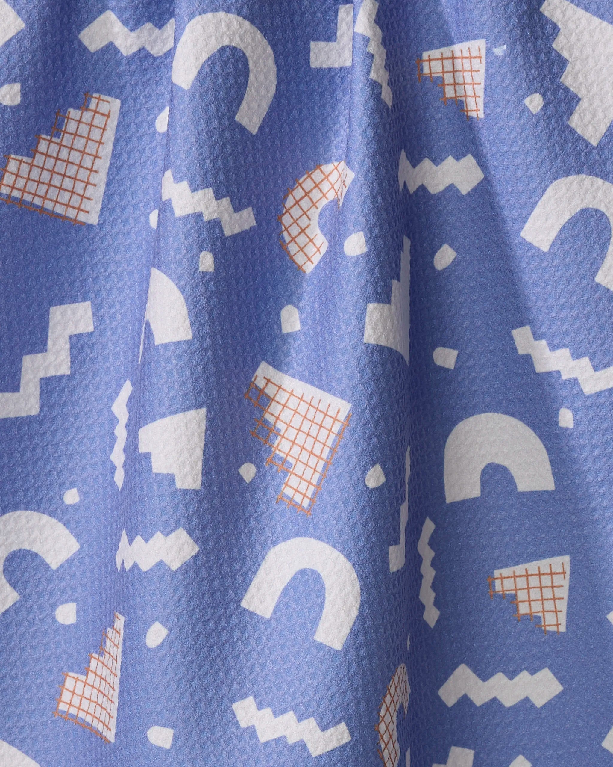 A close-up of Claire Iglesias’s Cutout Shapes fabric shows playful blue, white, and orange patterns on a textured surface—ideal for a vibrant, eco-friendly kitchen towel.