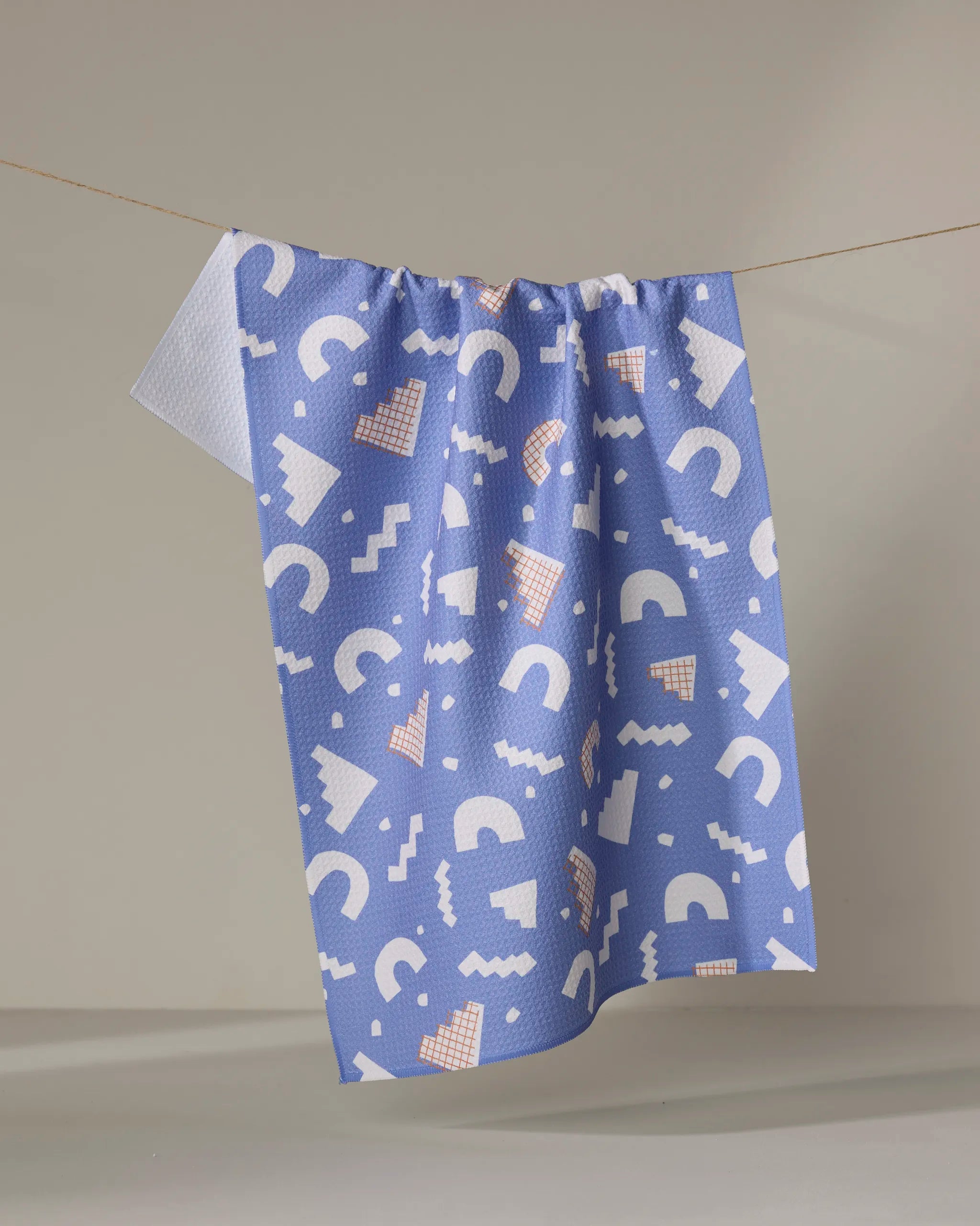 The Claire Iglesias "Cutout Shapes" eco-friendly kitchen towel in blue, featuring white and peach geometric patterns, hangs on a line against a plain light background.