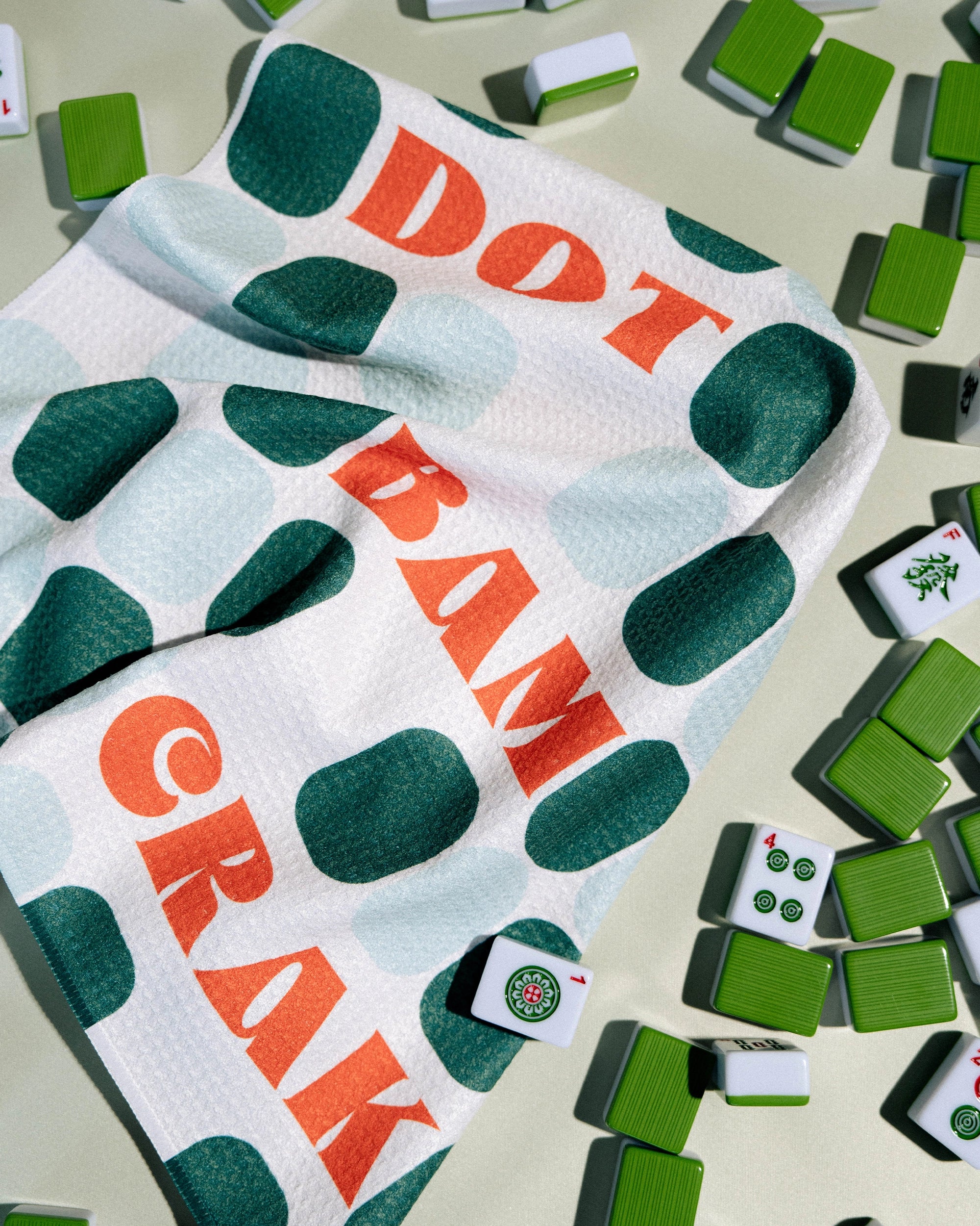 A Cypress Tile Talk recycled kitchen towel by Geometry, featuring green dots and the phrase "Dot Bam Crak," is draped among scattered mahjong tiles on a light surface.