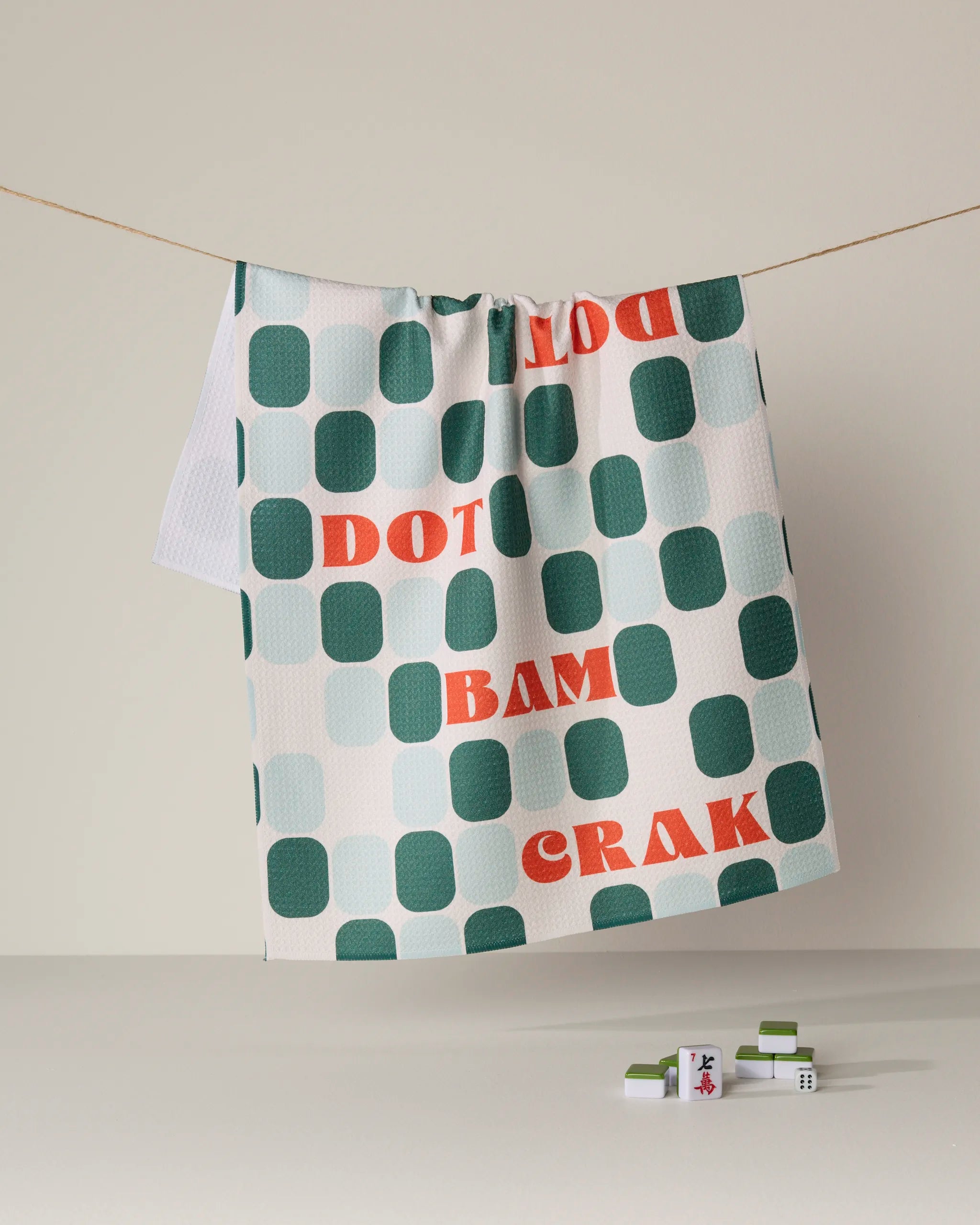 A white Cypress Tile Talk tea towel from Geometry, crafted from recycled Geoweave fabric, hangs on a line. It features teal squares, red "DOT BAM CRAK" text, and three Mahjong tiles displayed on a white surface below.