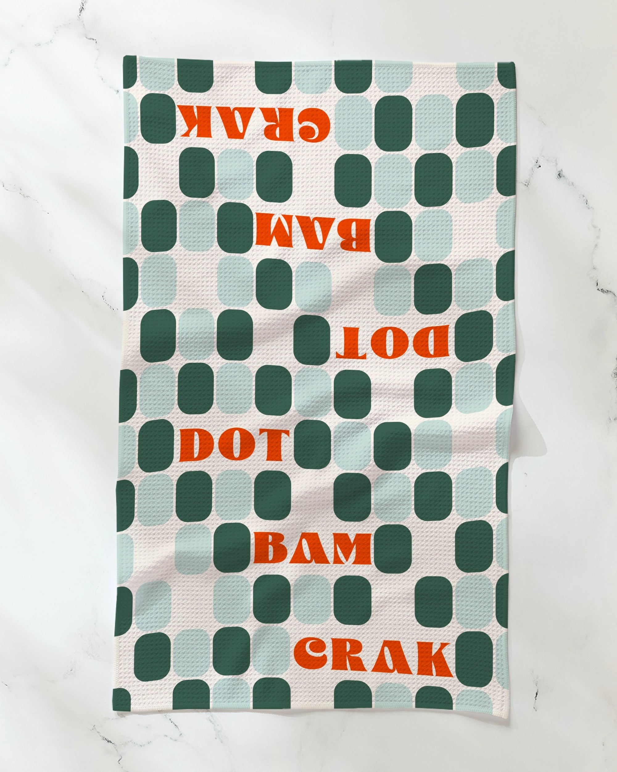 The Geometry Cypress Tile Talk dish towel is a patterned, absorbent towel featuring green and teal rounded squares with bold red "DOT BAM CRAK" lettering—some upside down—on a white background.