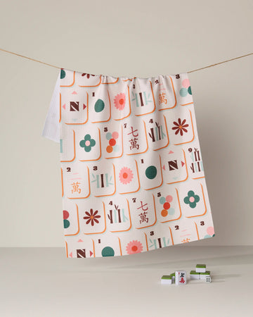 The Cypress Tile Toss kitchen tea towel by Geometry features vibrant, colorful mahjong tile patterns and hangs on a line with three tiles resting below on a neutral surface. Crafted from recycled materials, this ultra-absorbent towel combines the brand’s playful design with eco-friendly functionality.