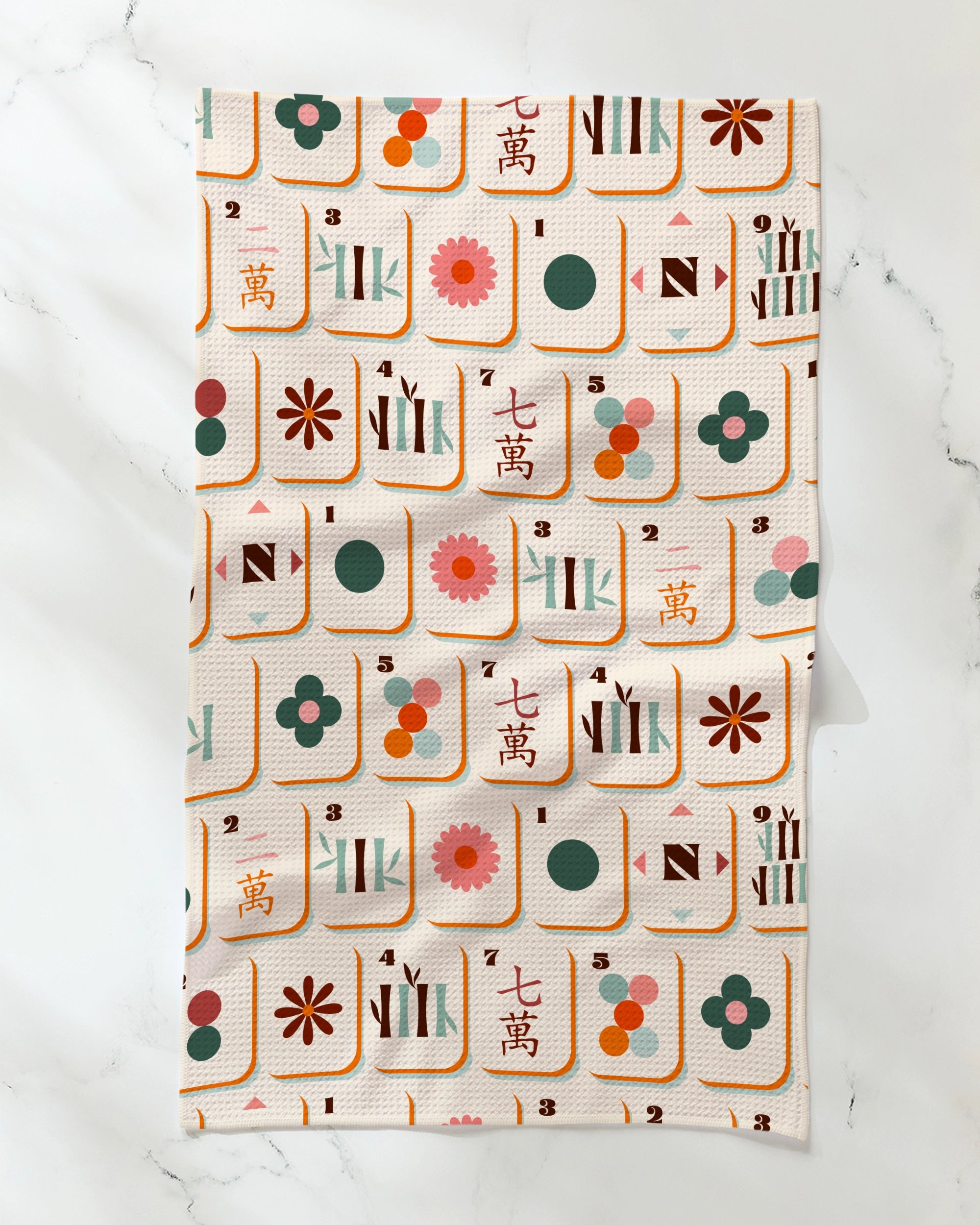 The Geometry Cypress Tile Toss tea towel showcases rows of stylized Mahjong tiles with floral and geometric patterns, beautifully displayed on a white marble surface. Crafted from recycled materials, this ultra-absorbent towel blends sustainability with stylish design.