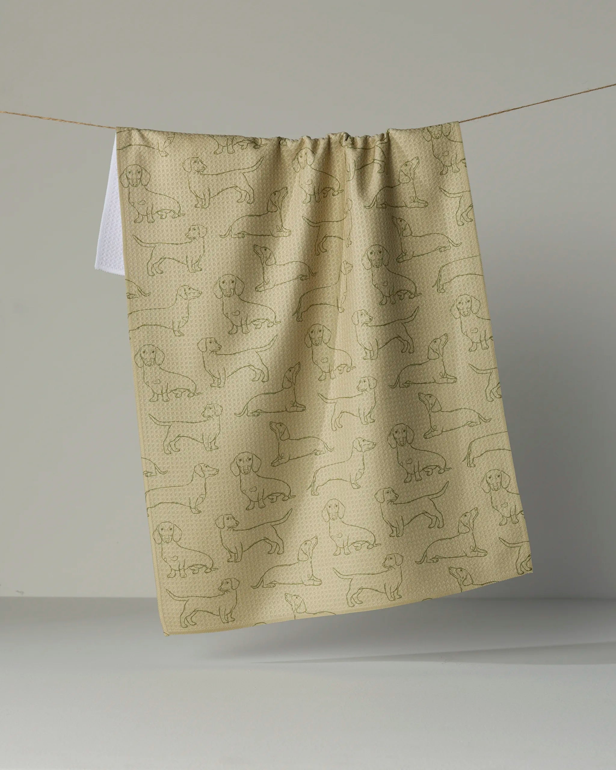 A “Dachshund Pups” tea towel by Geometry, featuring a pattern of outlined dachshund dogs and crafted from recycled materials in beige, hangs on a clothesline against a plain background.