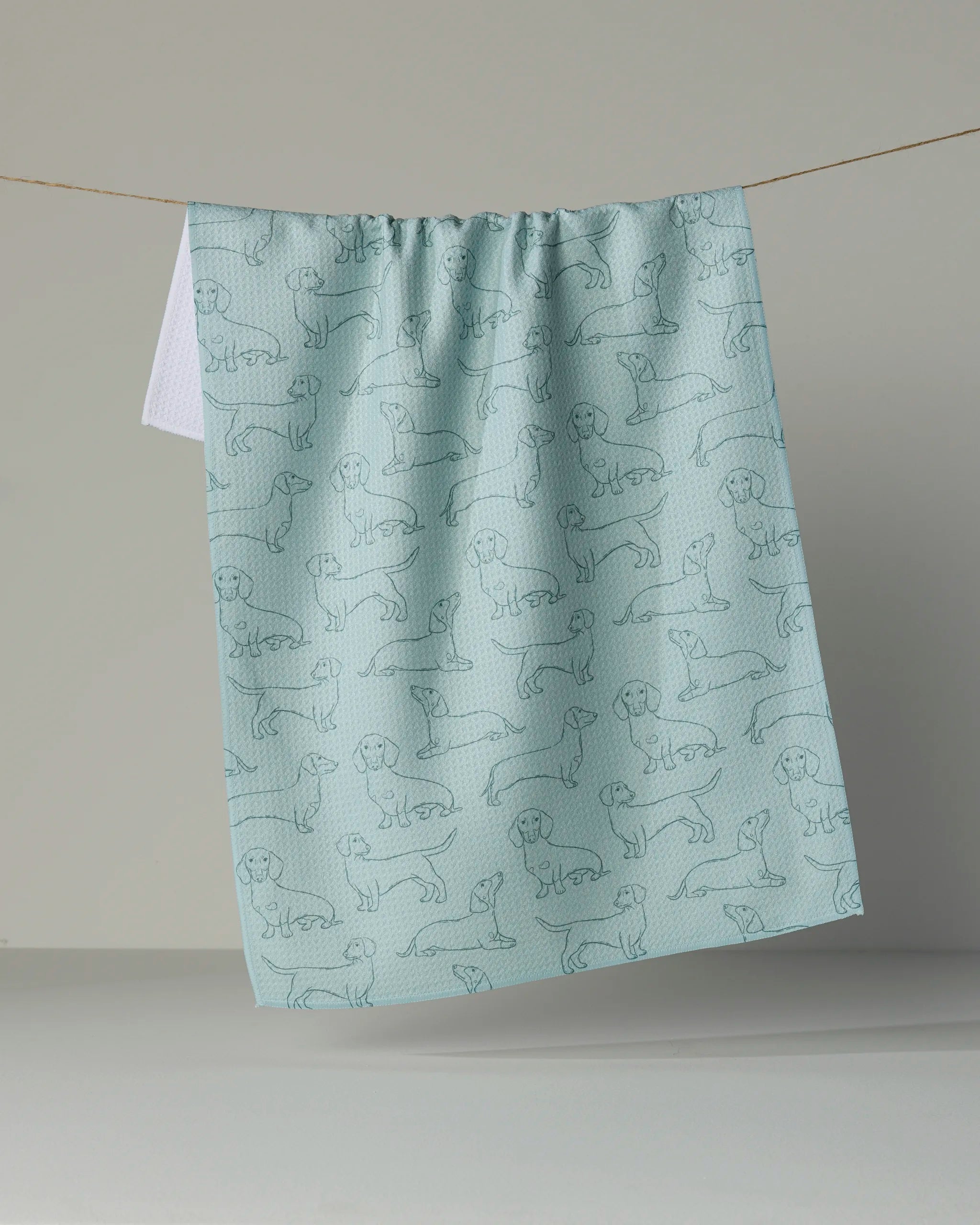A light blue Geometry kitchen towel, called “Dachshund Pups,” hangs on a line and features a pattern of simple, outlined dachshund dog illustrations.