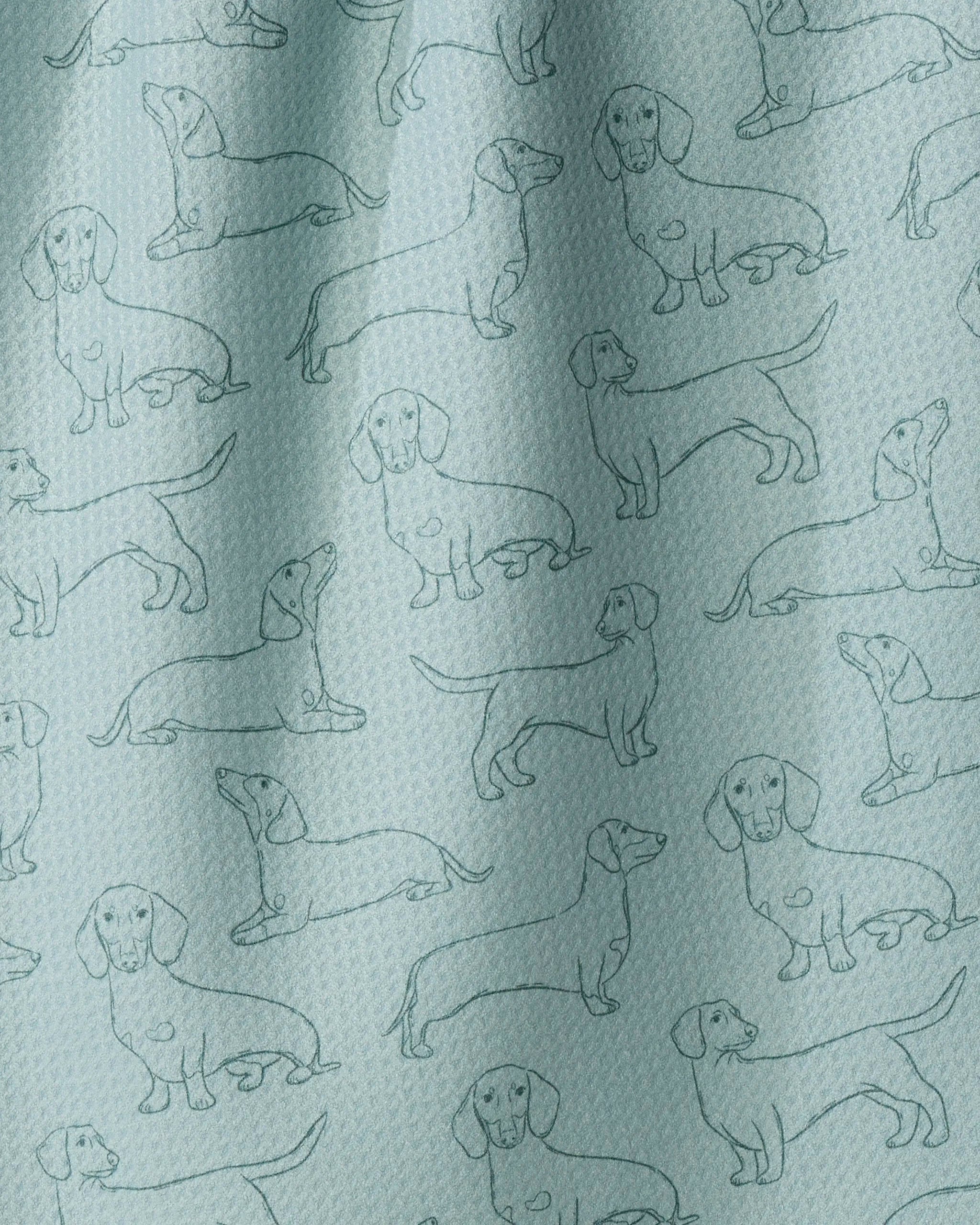 Geometry’s “Dachshund Pups” kitchen towel, crafted from recycled materials in a light blue hue, features a repeated line drawing pattern of dachshund dogs in various poses. The textured fabric displays gentle folds and subtle draping.
