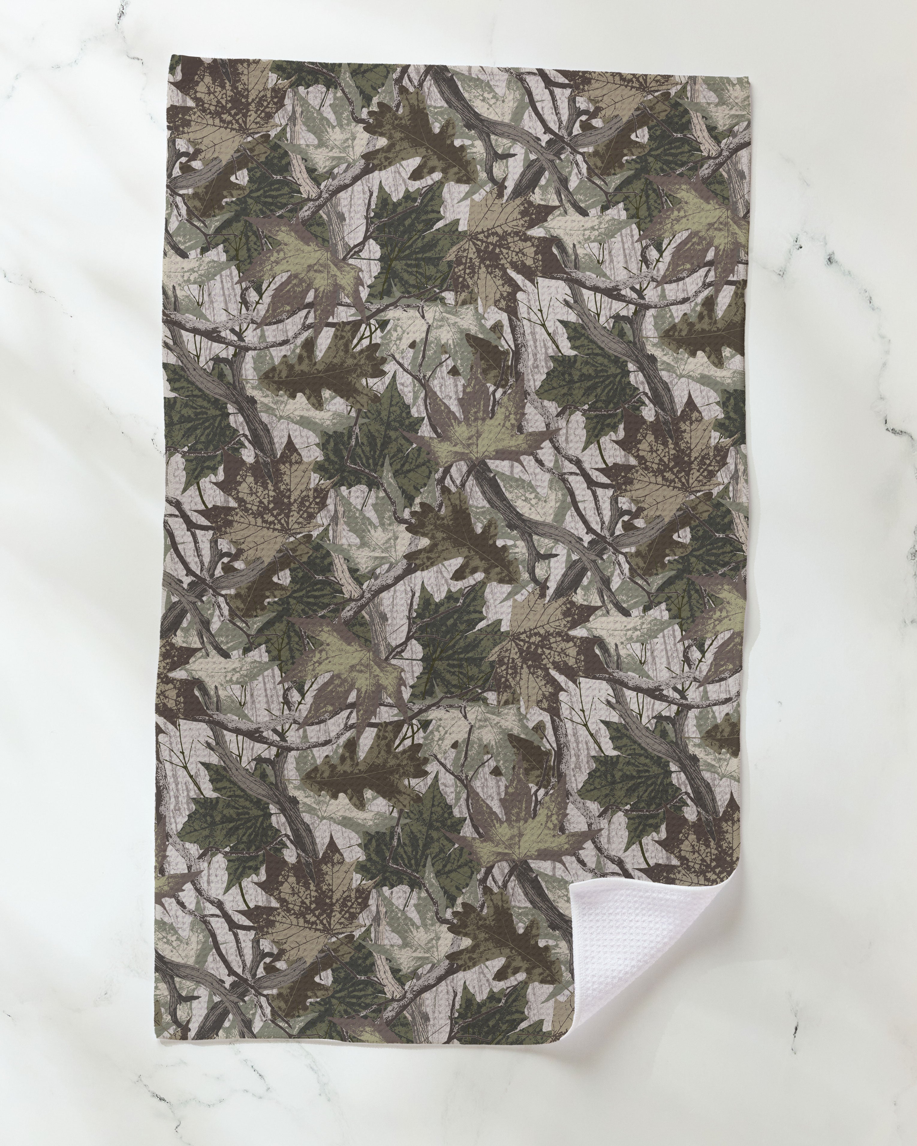 The Geometry "Dad Camo" kitchen towel, featuring a green and brown camouflage leaf pattern, lies flat on a white marble surface with one corner turned up to reveal its white textured underside crafted from recycled materials.