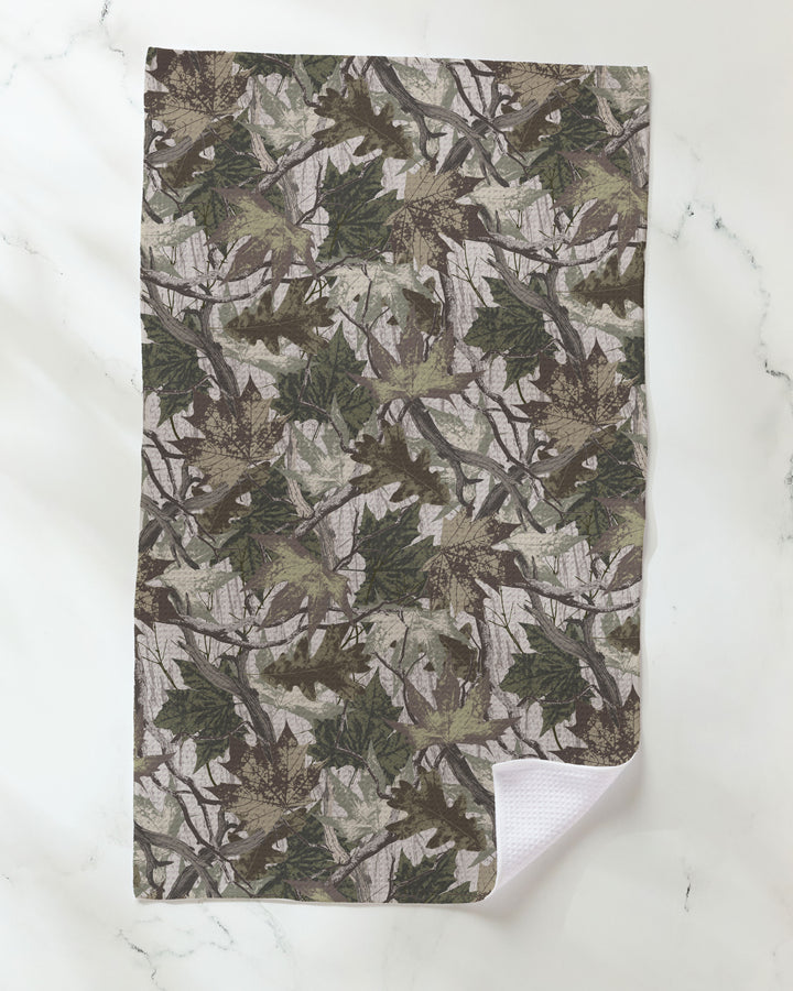 The Geometry "Dad Camo" kitchen towel, featuring a green and brown camouflage leaf pattern, lies flat on a white marble surface with one corner turned up to reveal its white textured underside crafted from recycled materials.