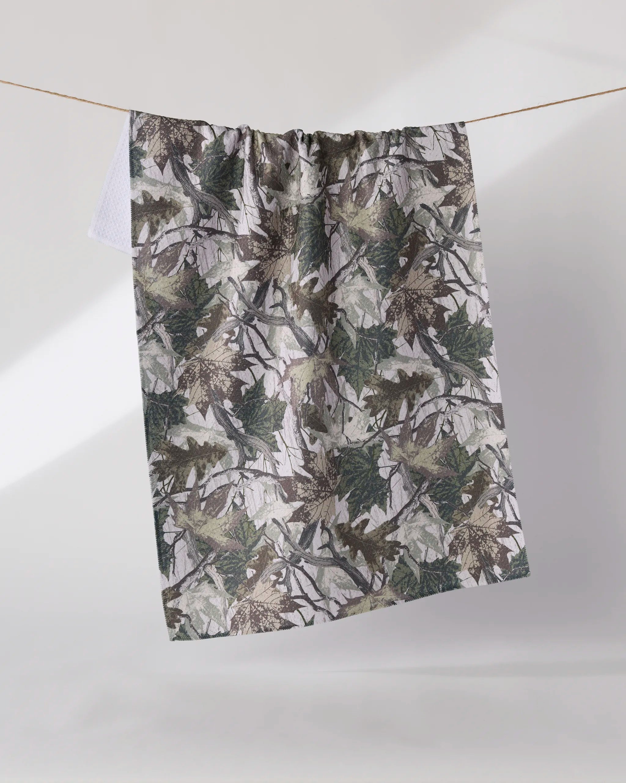 The Geometry Dad Camo kitchen tea towel, featuring a green and brown camouflage leaf pattern, hangs on a clothesline against a plain light background.