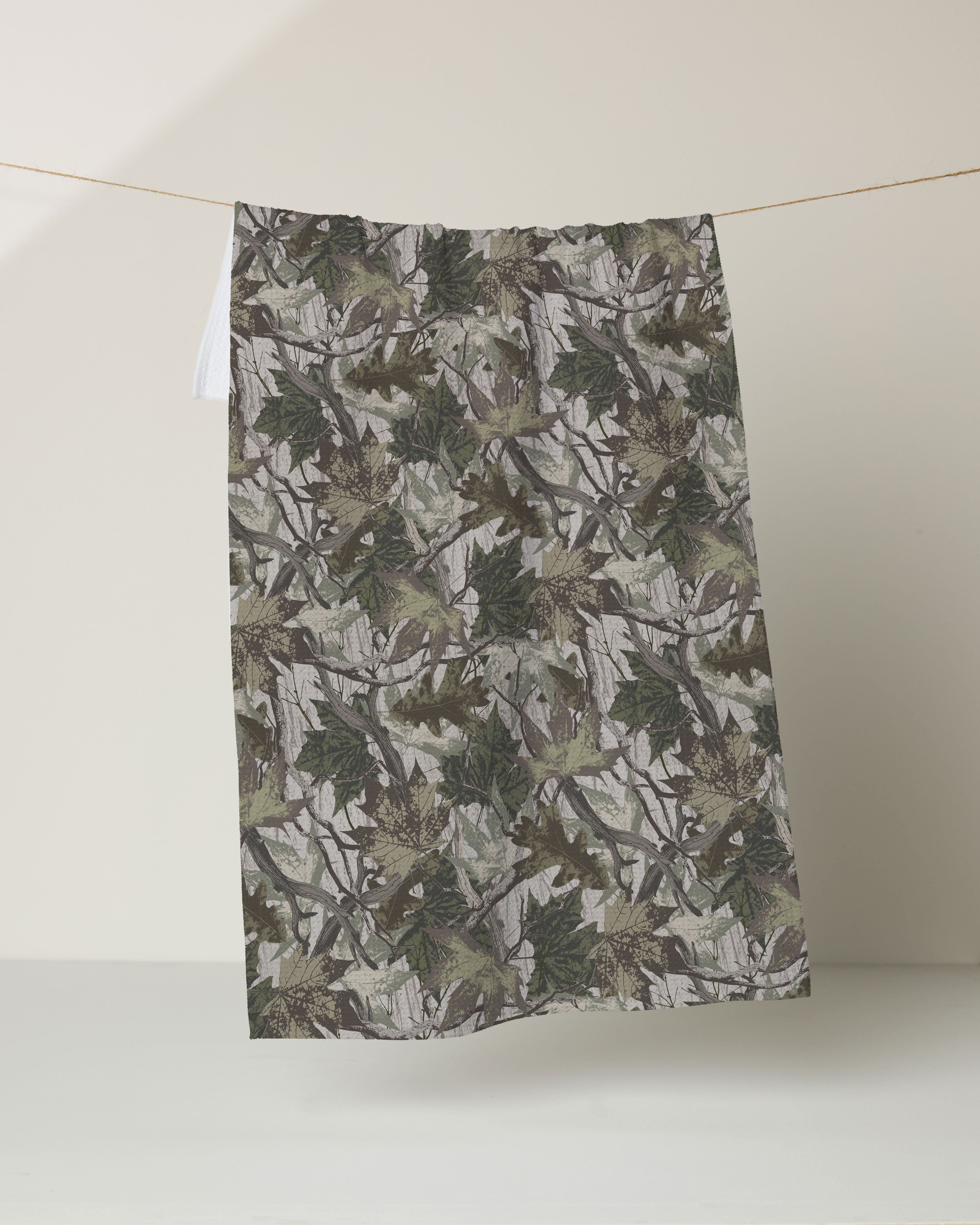 The Geometry Dad Camo kitchen towel, featuring a green and brown camouflage leaf design and made from recycled materials, hangs from a thin string against a plain, light-colored background.
