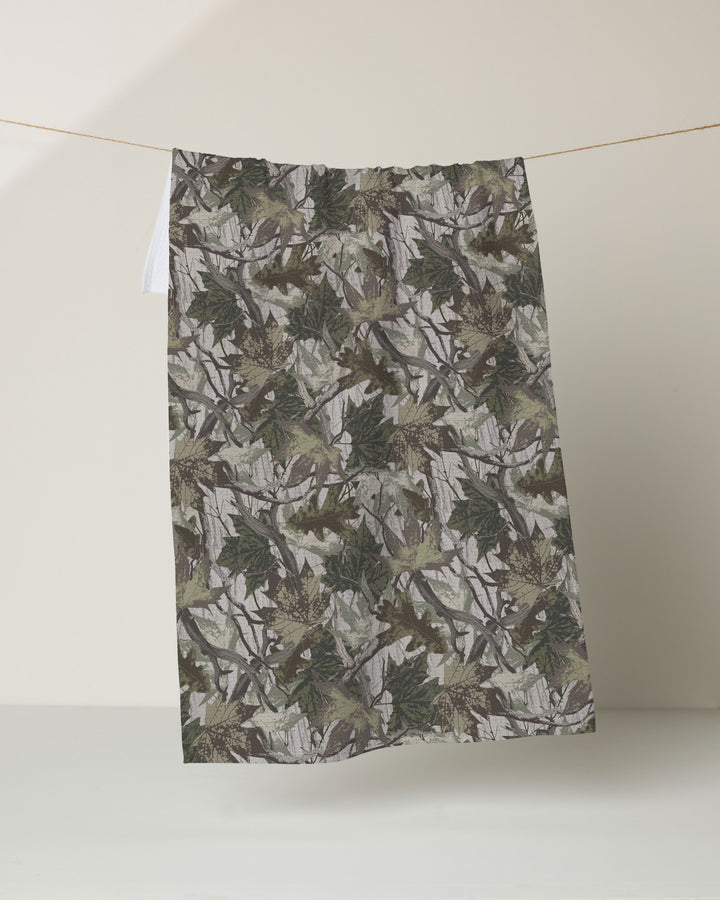 The Geometry Dad Camo kitchen towel, featuring a green and brown camouflage leaf design and made from recycled materials, hangs from a thin string against a plain, light-colored background.